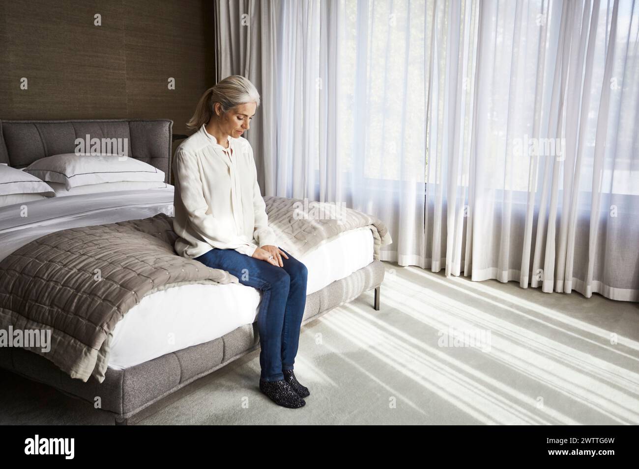 Woman sitting pensively on a bed in a well-lit room Stock Photo