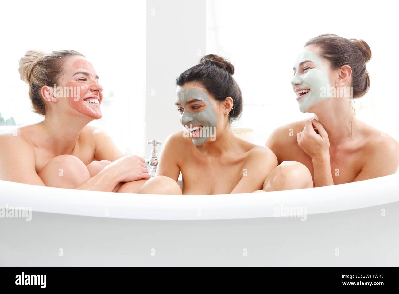 Three friends with facial masks laughing in a bathtub Stock Photo