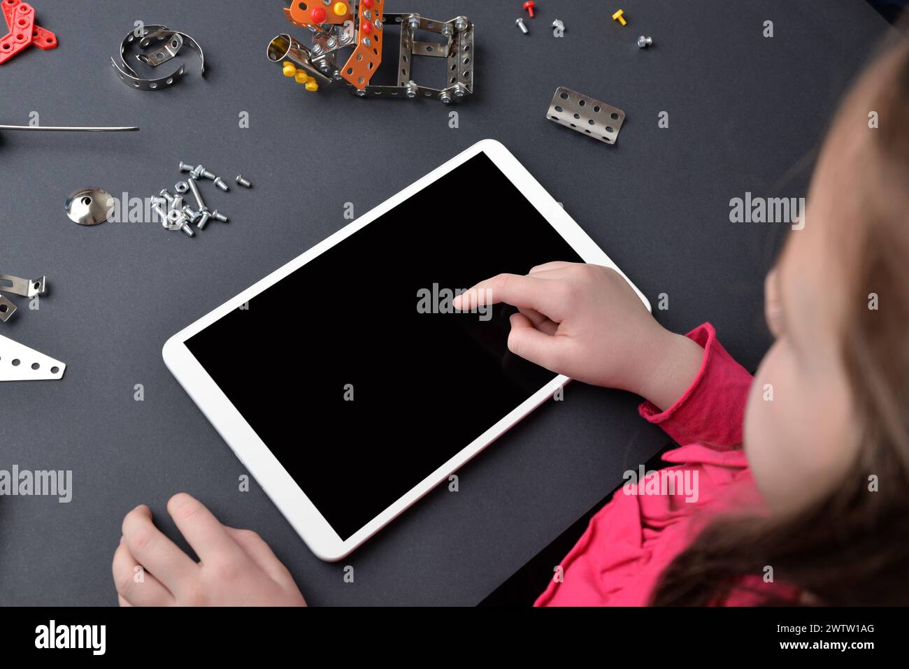 Little girl uses a tablet on a desk, surrounded by small parts and screws. The concept of education and composition of subjects, programming for child Stock Photo