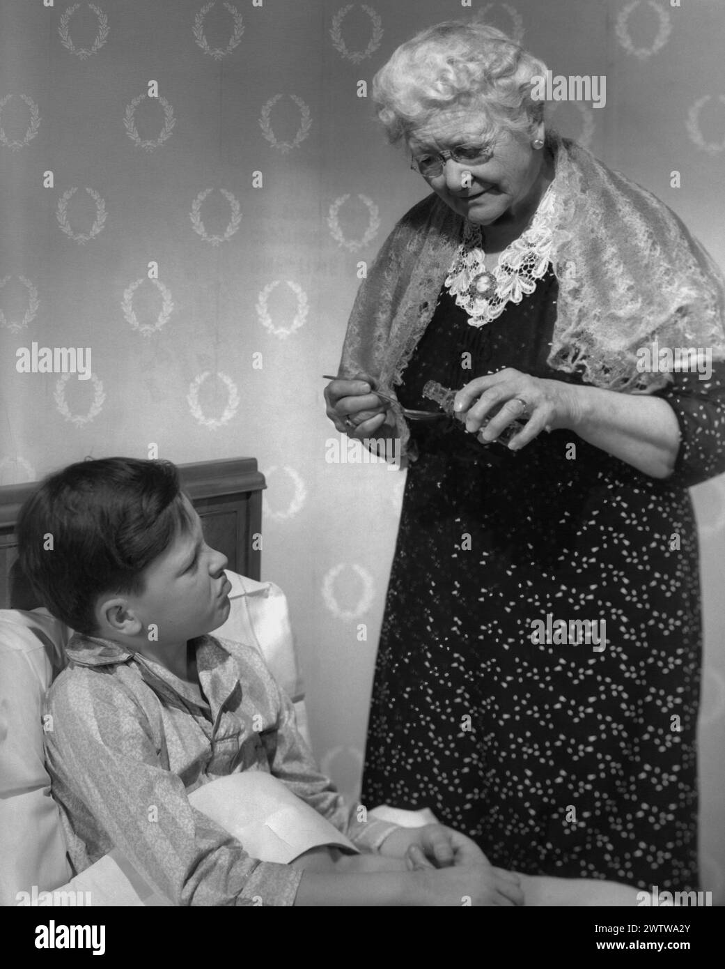 Older white-haired grandma with a dress and a shawl over her shoulders, standing bedside her grandson as she pours liquid medicine from a bottle into the spoon for him Stock Photo