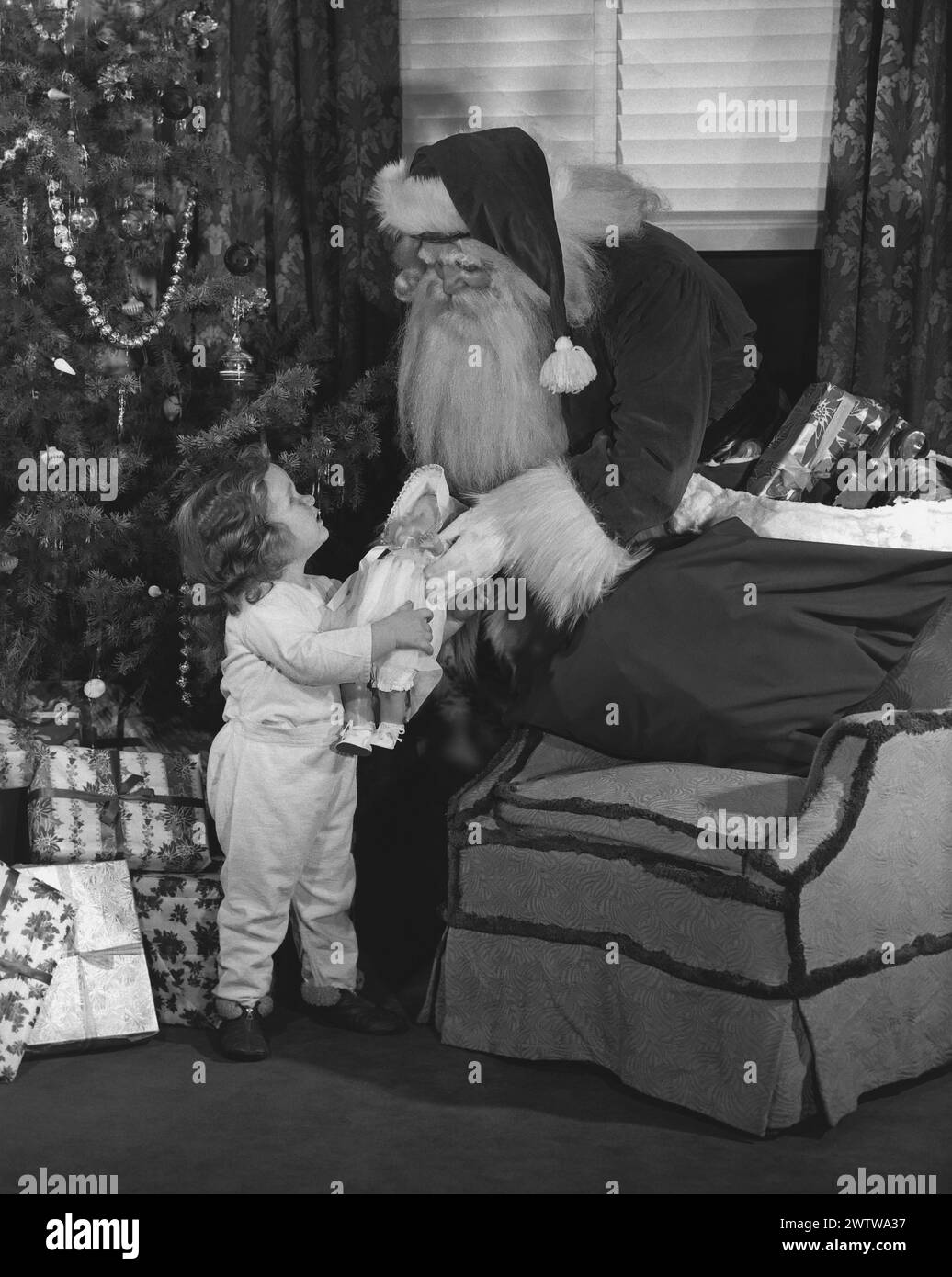 Santa Claus standing between the tree and a chair with his bag of toys on the chair handing over a new doll to a little girl Stock Photo