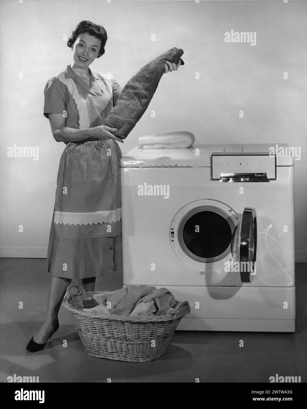 Young woman in a dress and a house apron as well as high heels, standing in front of her electric dryer with a basket of clothes on the floor holding a folded towel and smiling at the camera Stock Photo