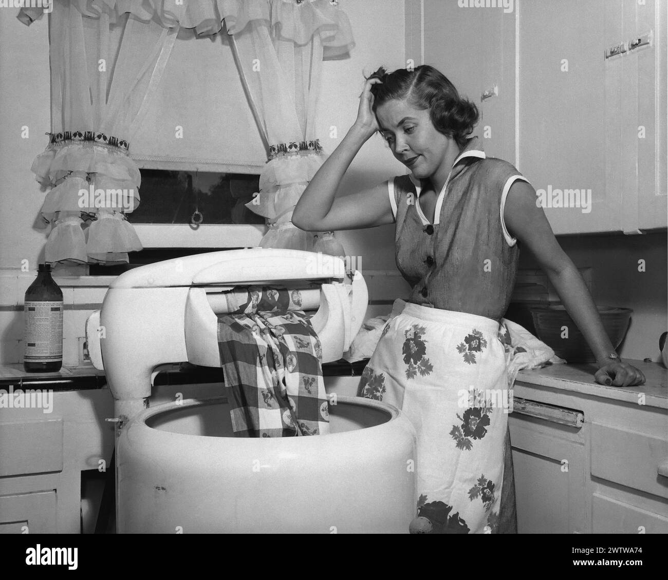 Young woman, well-dressed, with an apron on standing her kitchen looking exhausted while taking a break from washing laundry in a manual washtub Stock Photo