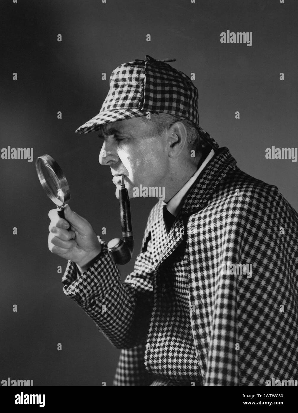 Older man in a Sherlock Holmes type matching outfit and hat with a hand and a magnifying glass up to his face with a large pipe hanging out of his mouth Stock Photo