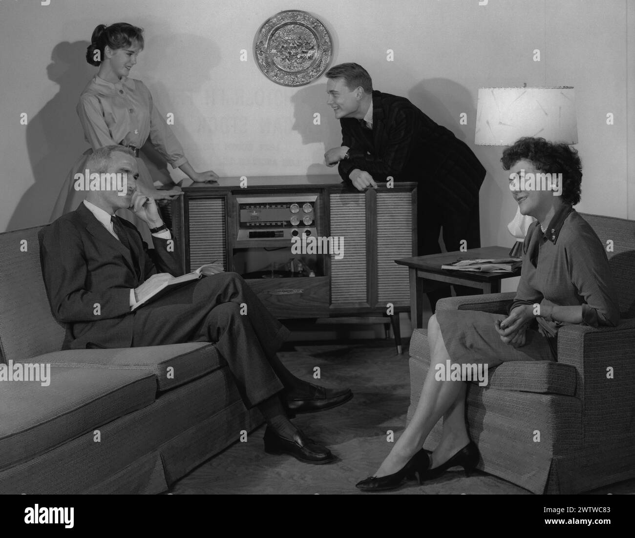 Family sitting in the living room, all well dressed, mother and father sit whilst their two grown children stand in the background, leaning on a phonograph cabinet Stock Photo