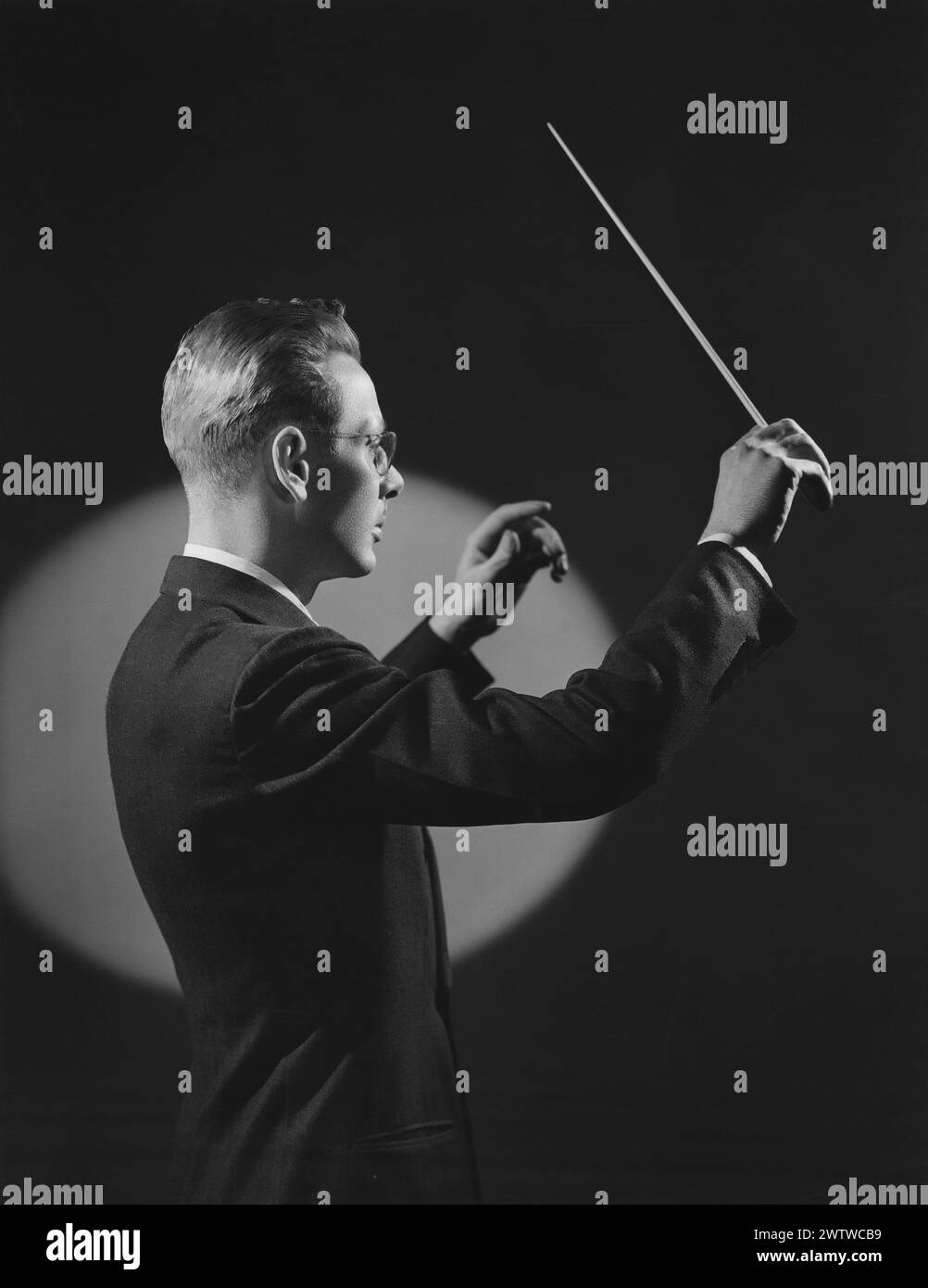 Dramatically lit male orchestra conductor shot from the side with a spotlight lighting him and the background while he has his hands in the air and waving a baton. Stock Photo