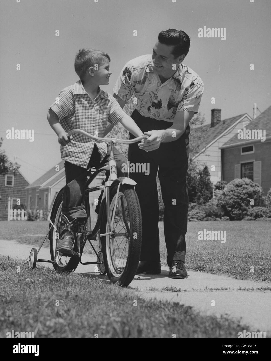 Dad and son pause to look at each other while learning to ride a bicycle on the sidewalk in a