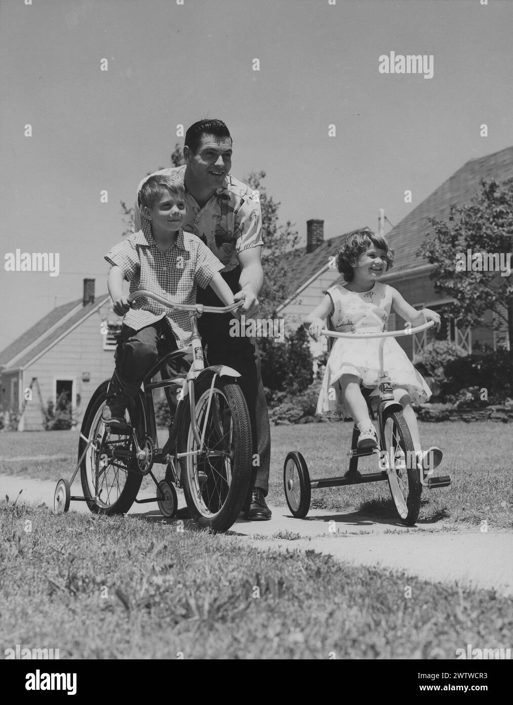 Young, a father helping his son learn how to ride a two wheel bike with training wheels down the sidewalk as his daughter writes a tricycle next to them. All are looking ahead. Stock Photo