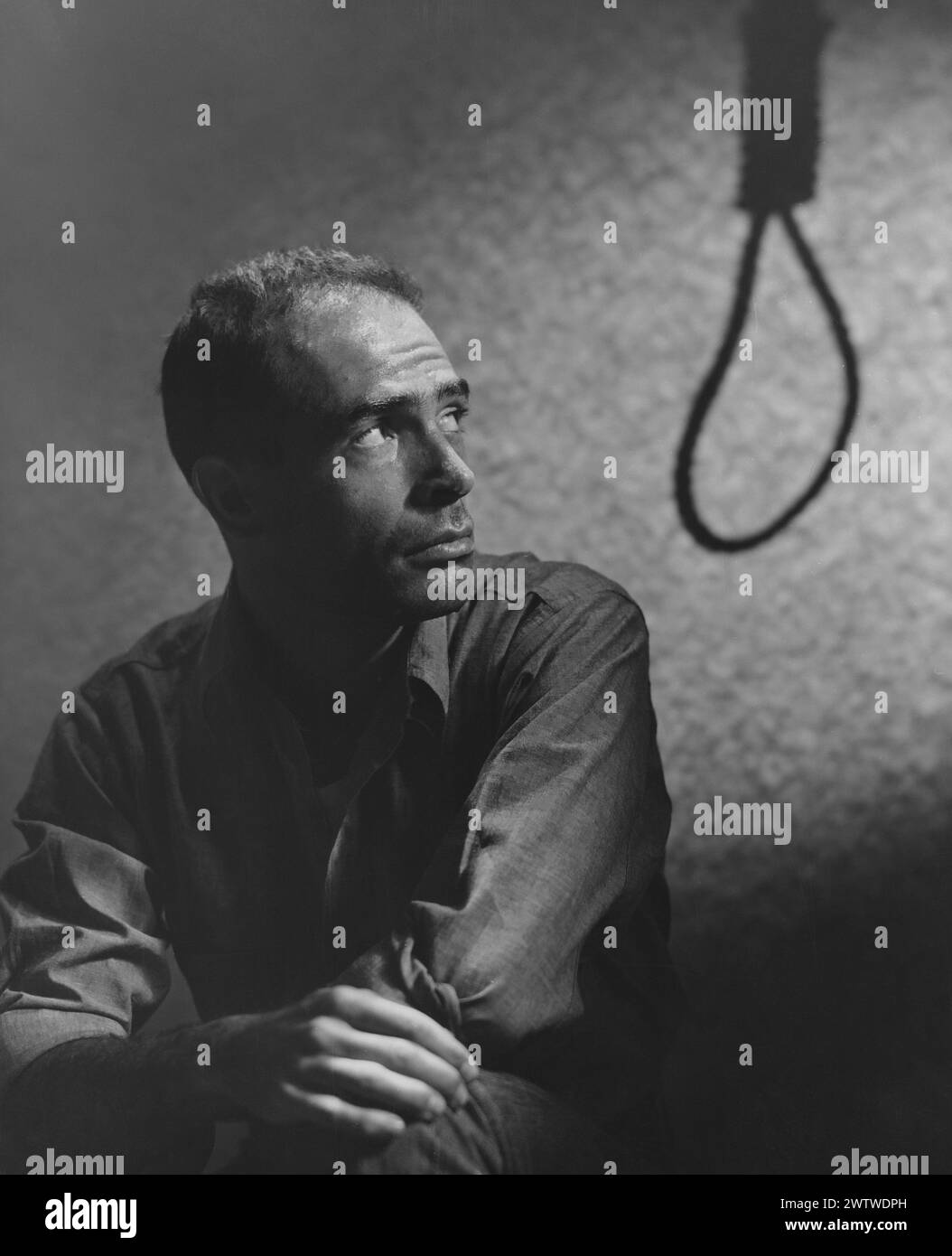 Middle-aged white man sitting in a dimly lit room, looking back over his shoulder at a silhouette of a noose Stock Photo