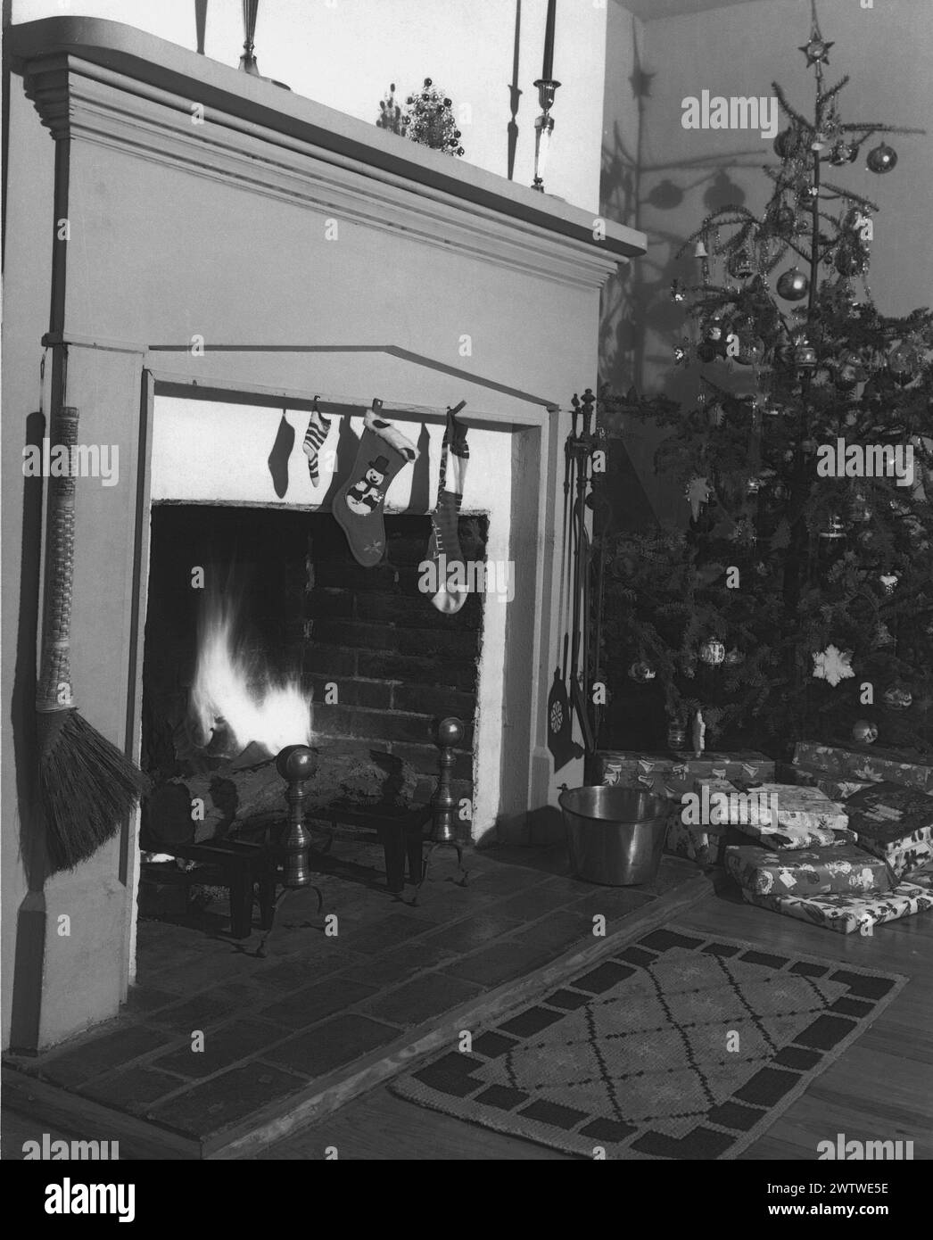 blazing fire in the grate of a decorated fireplace next to a Christmas tree which has presents underneath Stock Photo