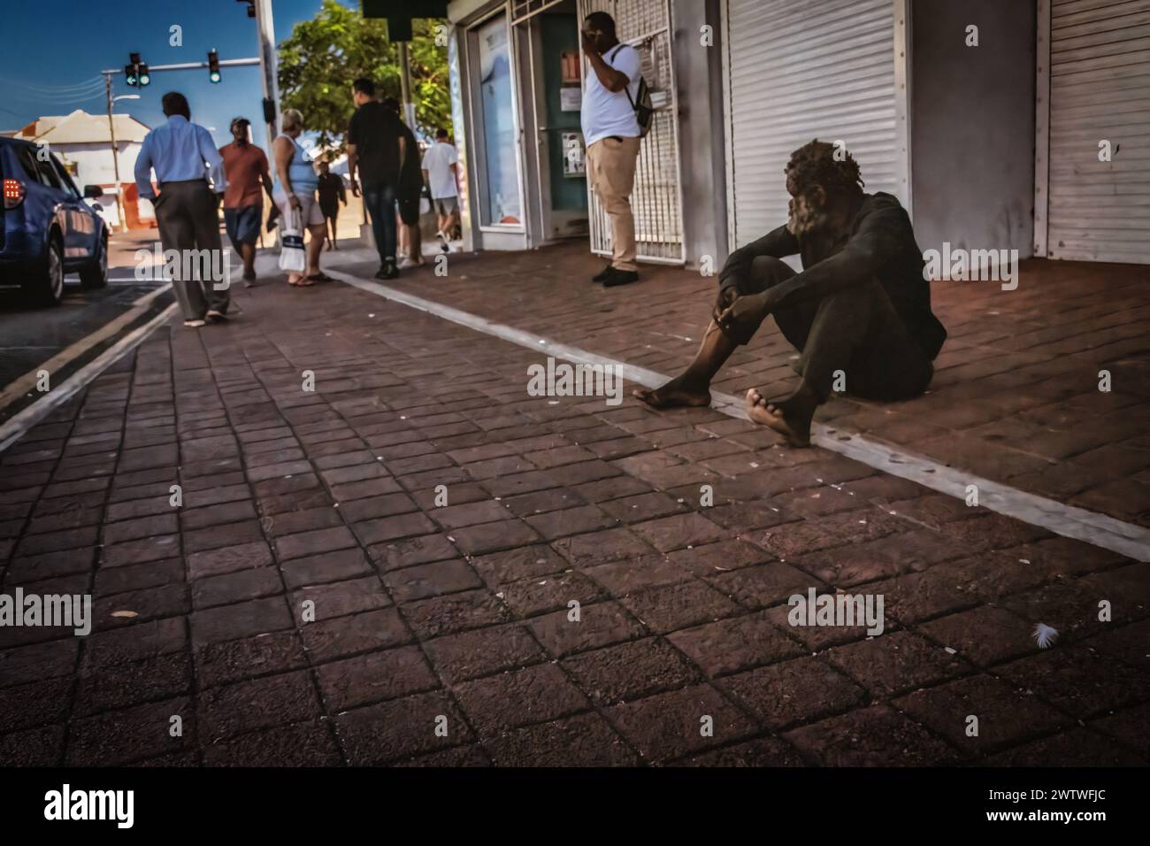 Depicting isolation, a lone person sits on the sidewalk with their head down, while others pass by without notice Stock Photo