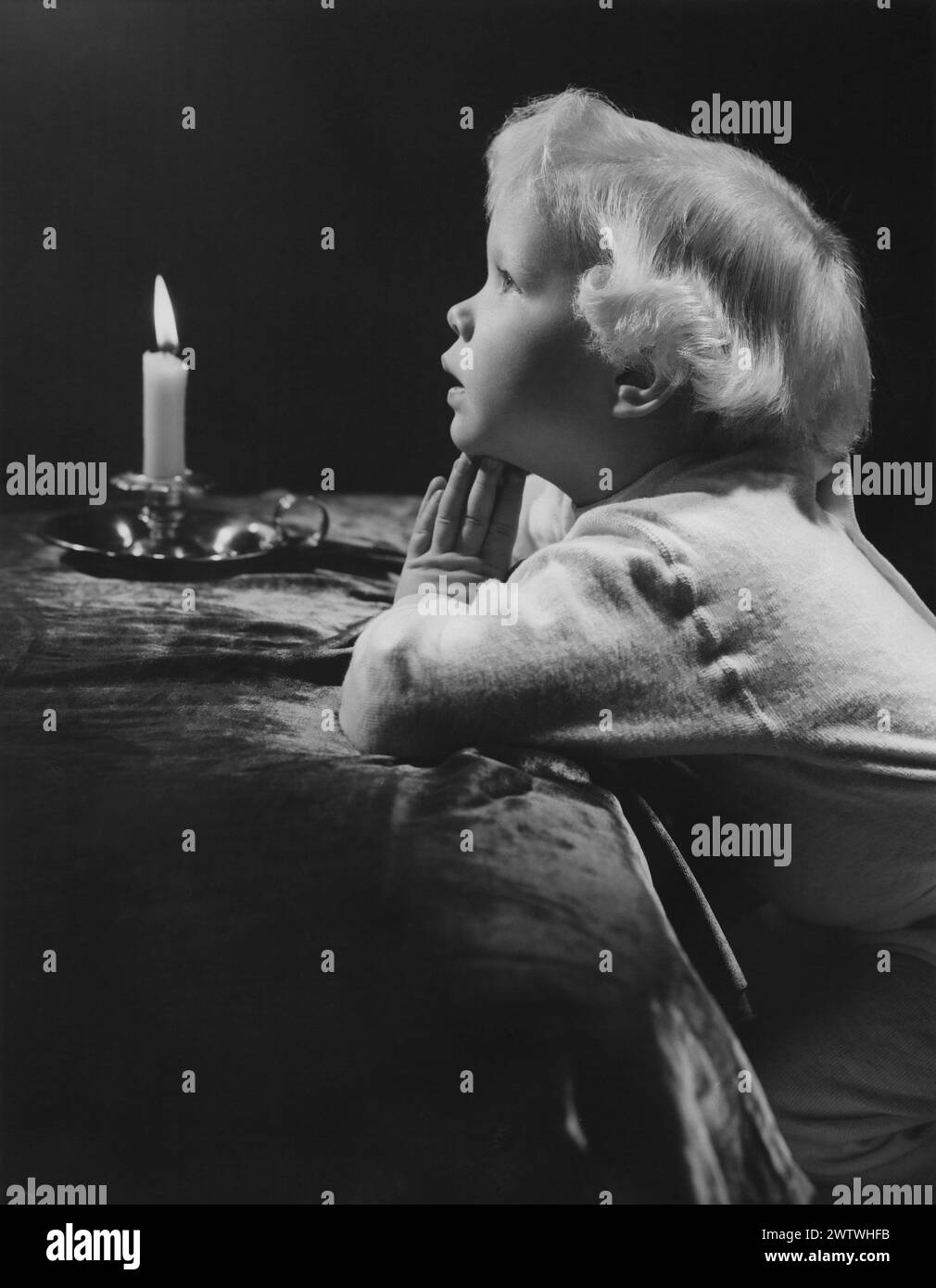 Blond headed boy kneeling next to his bed with hands clasped like he's praying, glancing upward with a candle burning in the background Stock Photo