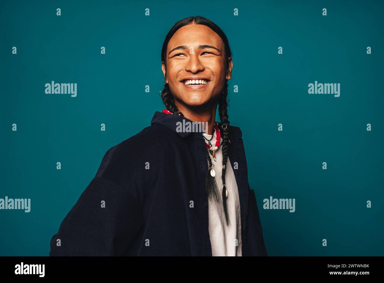 Confident, cheerful and stylish young man with braided hair stands against a blue background. Ethnic jewelry and casual clothing complement his unique Stock Photo