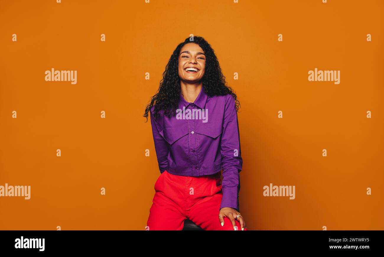 Cheerful woman with curly hair sits in casual clothing, smiling at the camera. Her vibrant, stylish fashion sense stands out against the orange backgr Stock Photo