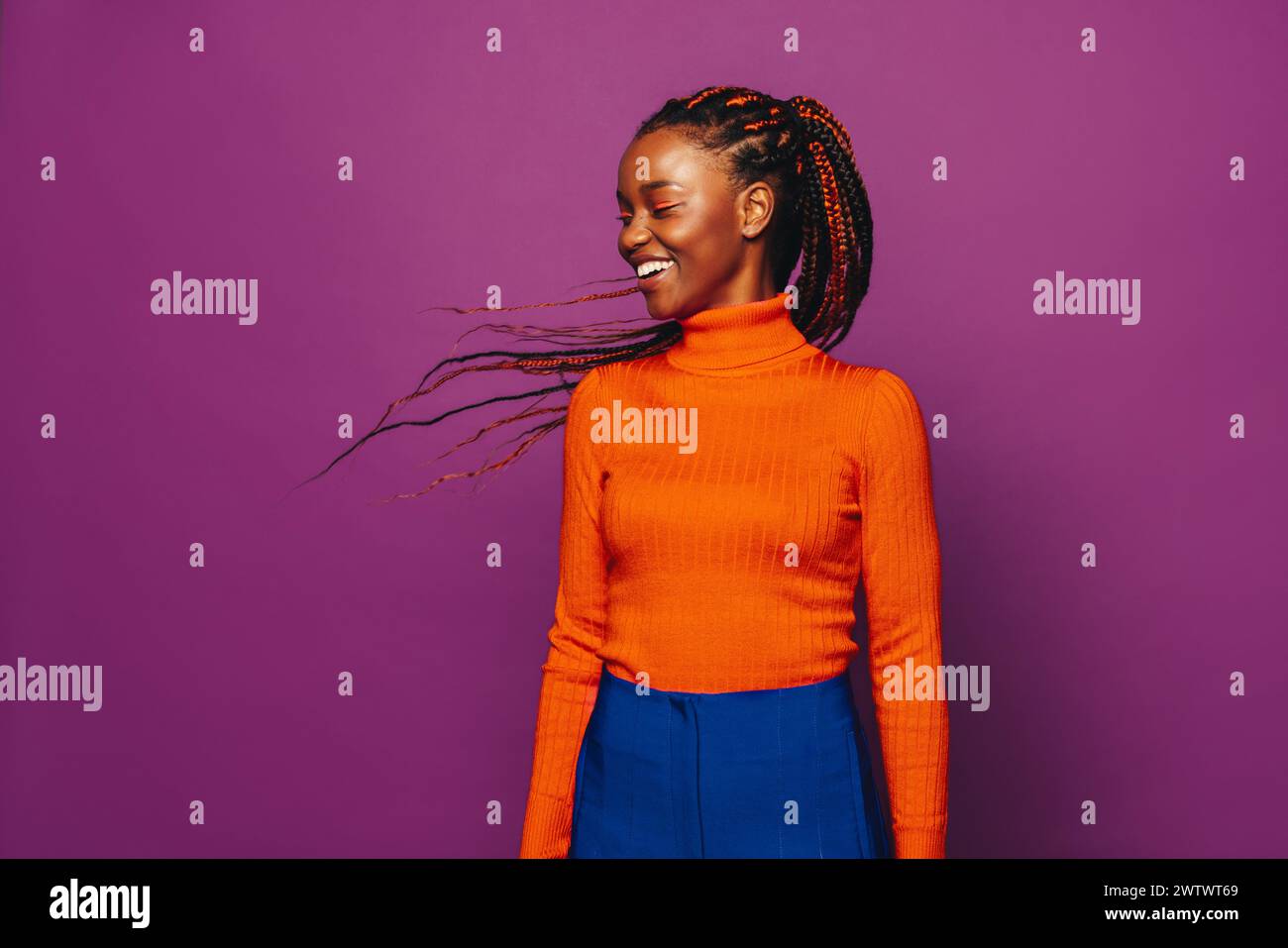 African Gen Z woman with stylish two-tone braids stands in a studio with a vibrant purple background. She wears casual clothing, including jeans and a Stock Photo