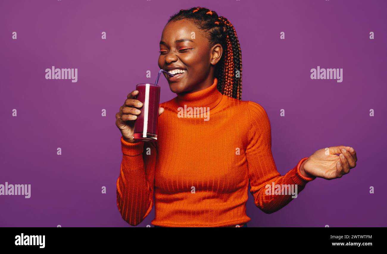 Happy woman with stylish braided hair stands against a purple background, holding a cup with a straw. She wears casual clothing and vibrant makeup, ra Stock Photo