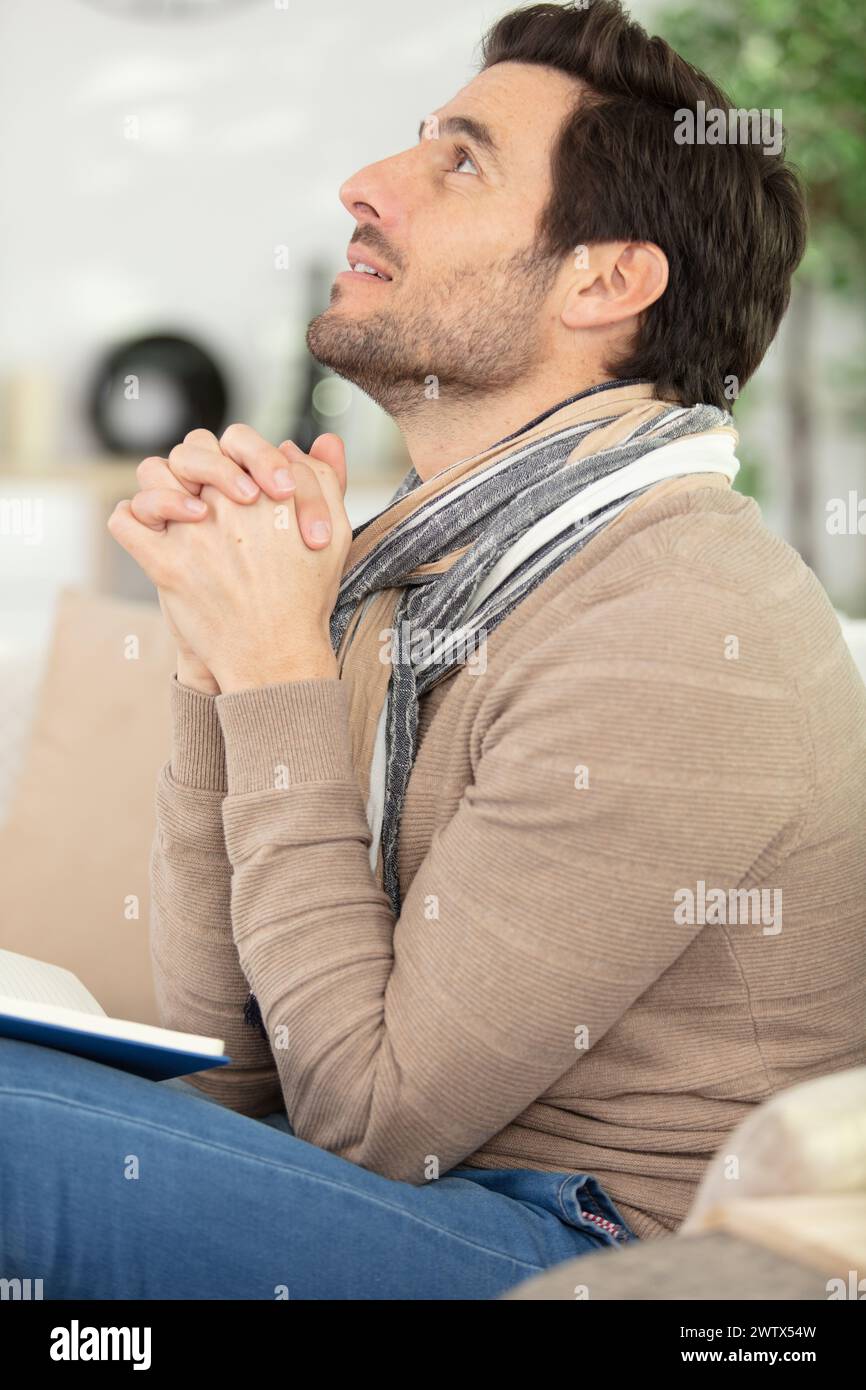 faithful middle aged man praying while sitting on sofa Stock Photo