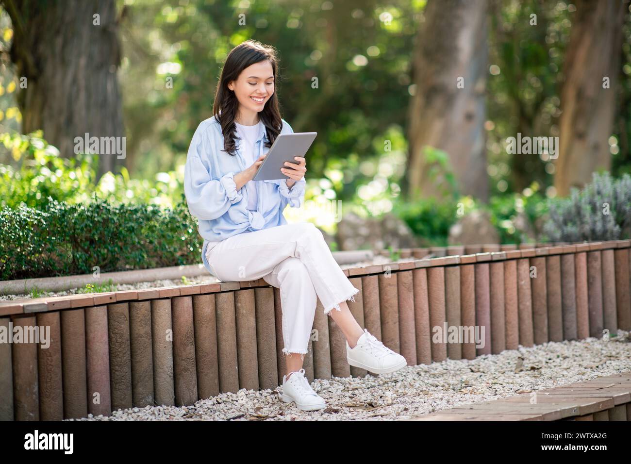 Engrossed young female student sitting cross-legged on a park bench, deeply focused on her tablet Stock Photo