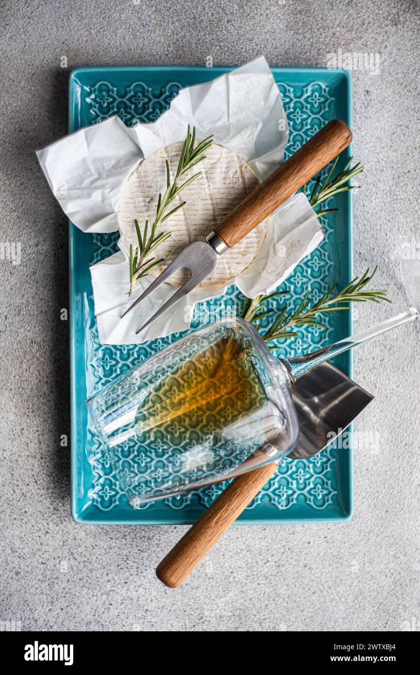 Overhead view of a whole brie with fresh rosemary on a plate with cheese knives and a glass of white wine Stock Photo