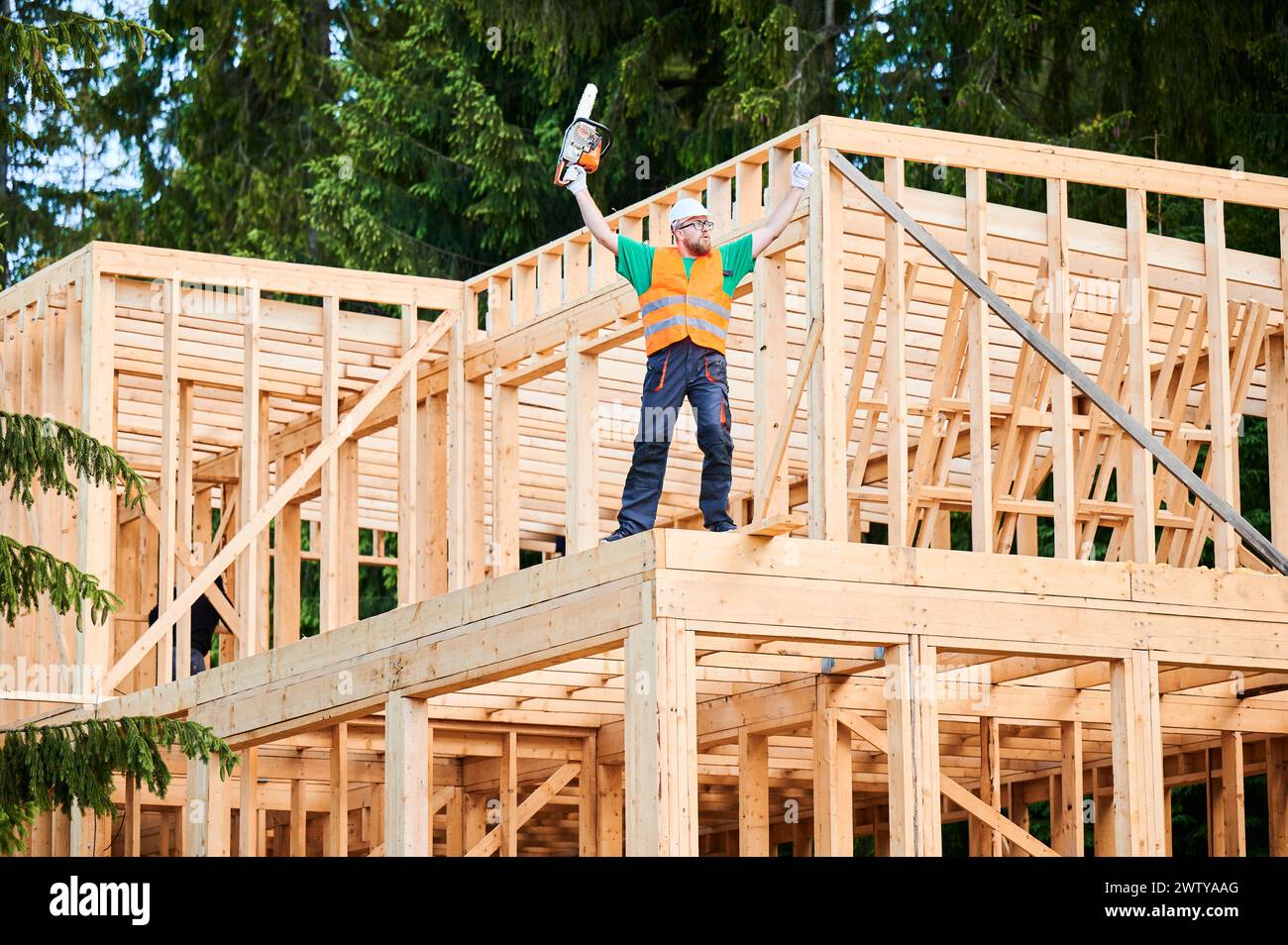 Carpenter working on construction site of wooden frame house. Man has calloused hands raised high and holds a chainsaw with which he processes the wooden beams for further use in construction. Stock Photo