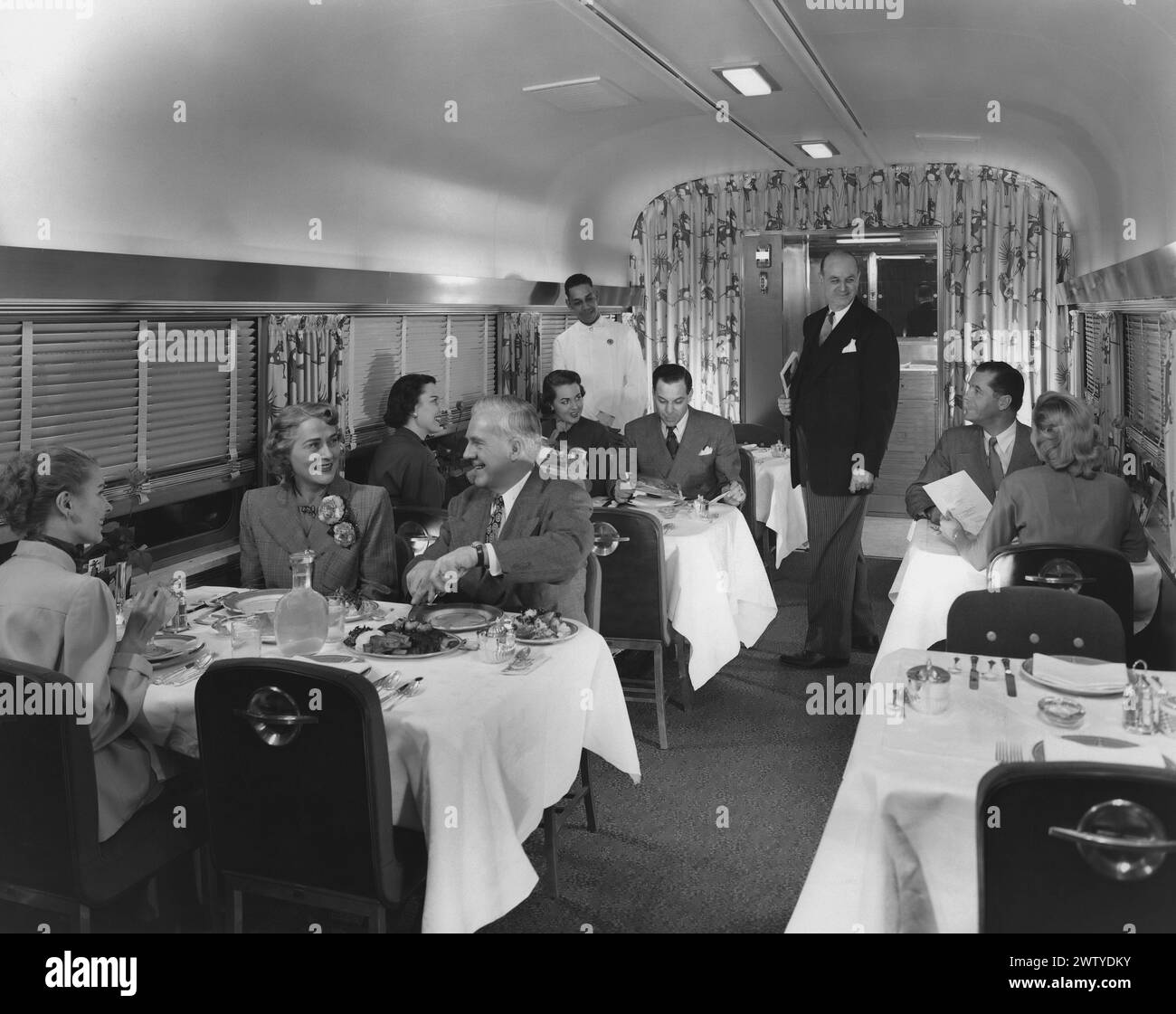 Well-dressed men and women sitting at white linen tablecloth covered tables with chinaware eating a meal in a railway car Stock Photo