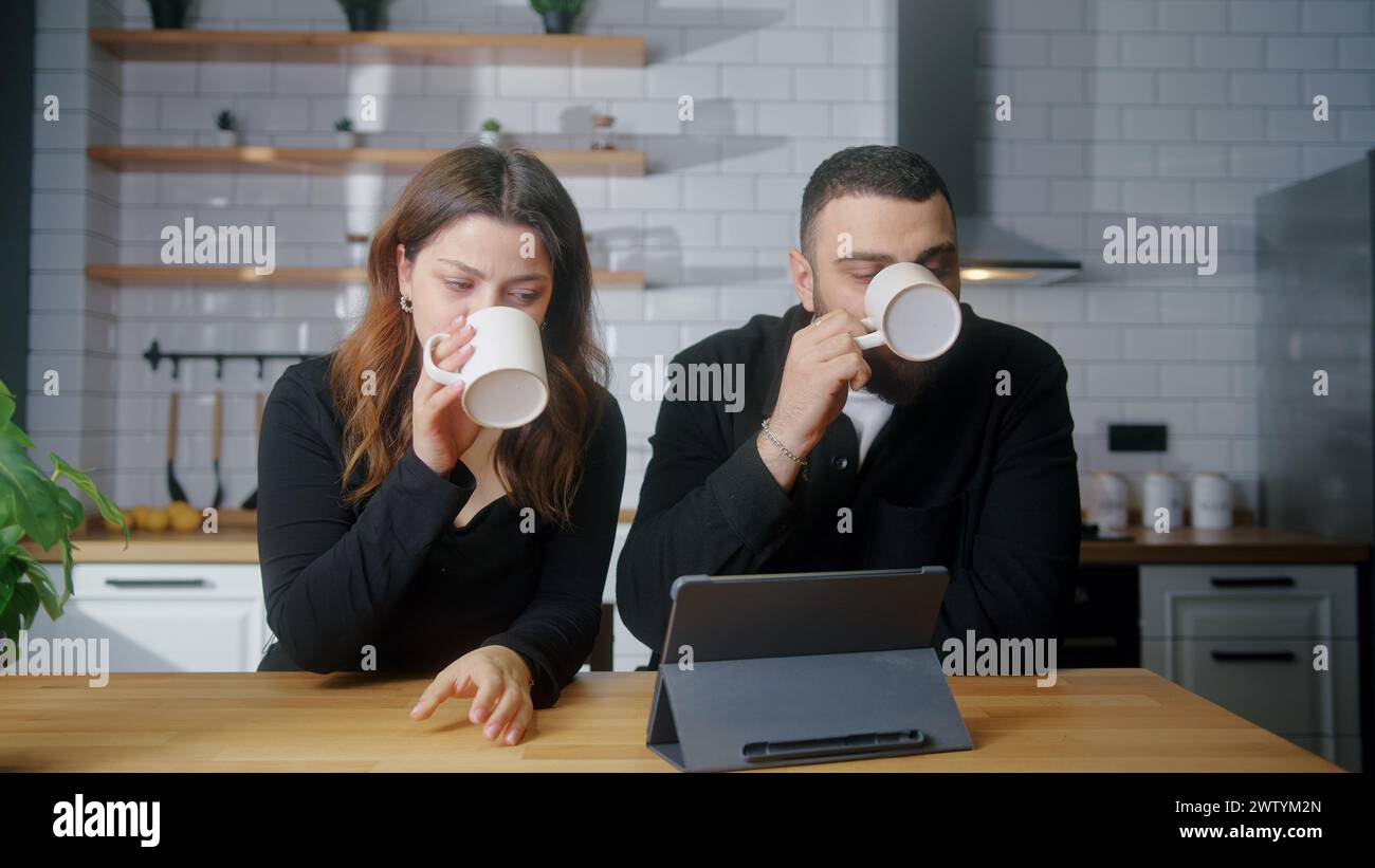 Young couple sit in kitchen use tablet computer, drinking tea or coffee, spend time together Stock Photo