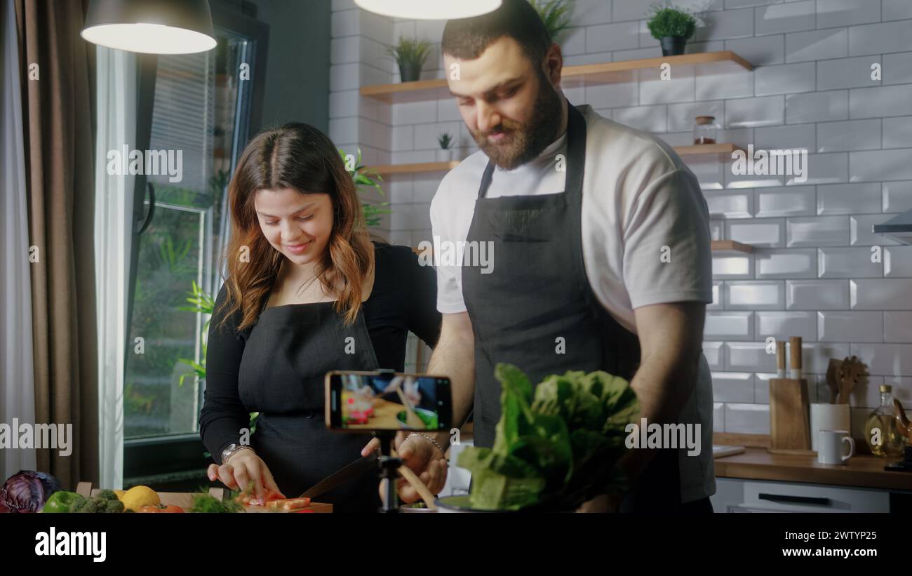 Young couple in an apron standing in kitchen records on smartphone food videoblog. Couple cutting vegetables, preparing healthy a salad. Blogging, hea Stock Photo