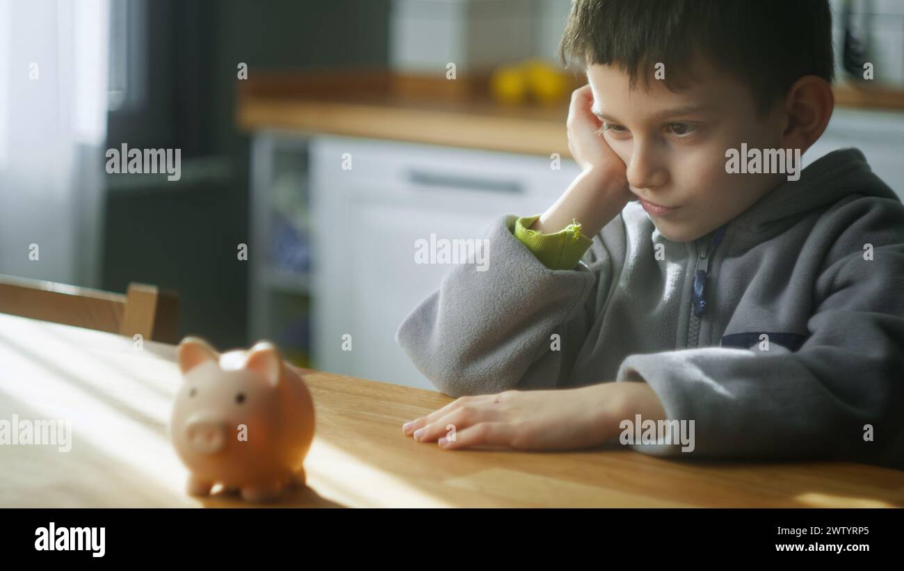 Bored cute boy sit in the kitchen thinking and looking to a piggy bank. Concept of teaching financial literacy, budget planning Stock Photo