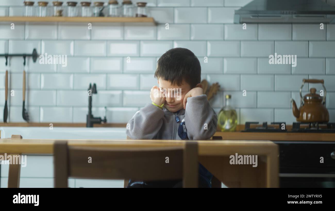 Little upset kid sitting in kitchen alone at home. Lonely offended schoolboy feeling disappointed bullied, stressed sit at table. School children prob Stock Photo