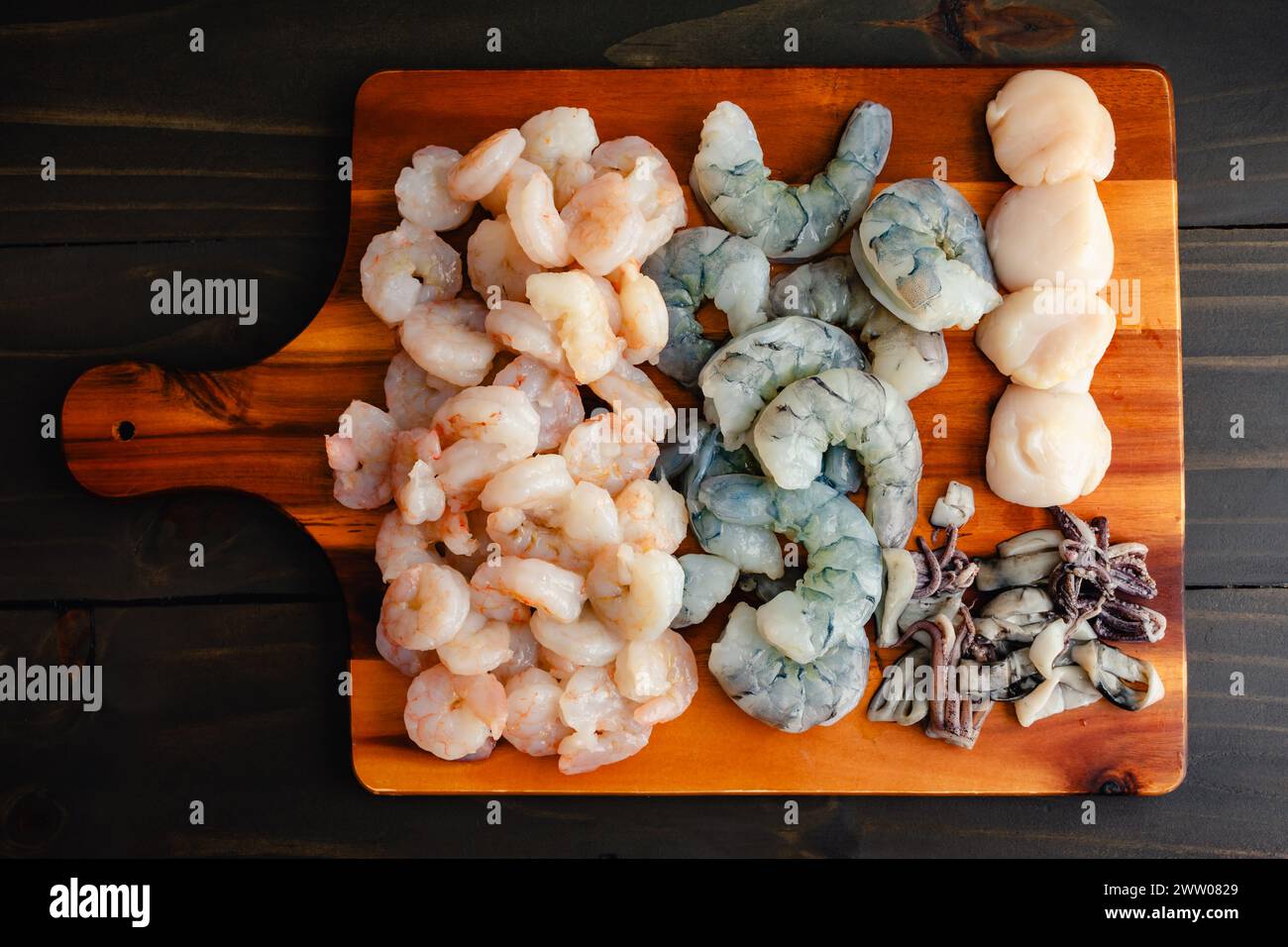 Raw Seafood Arranged on a Wooden Cutting Board: Uncooked peeled and deveined shrimp, sliced squid, and scallops on a wood chopping board Stock Photo