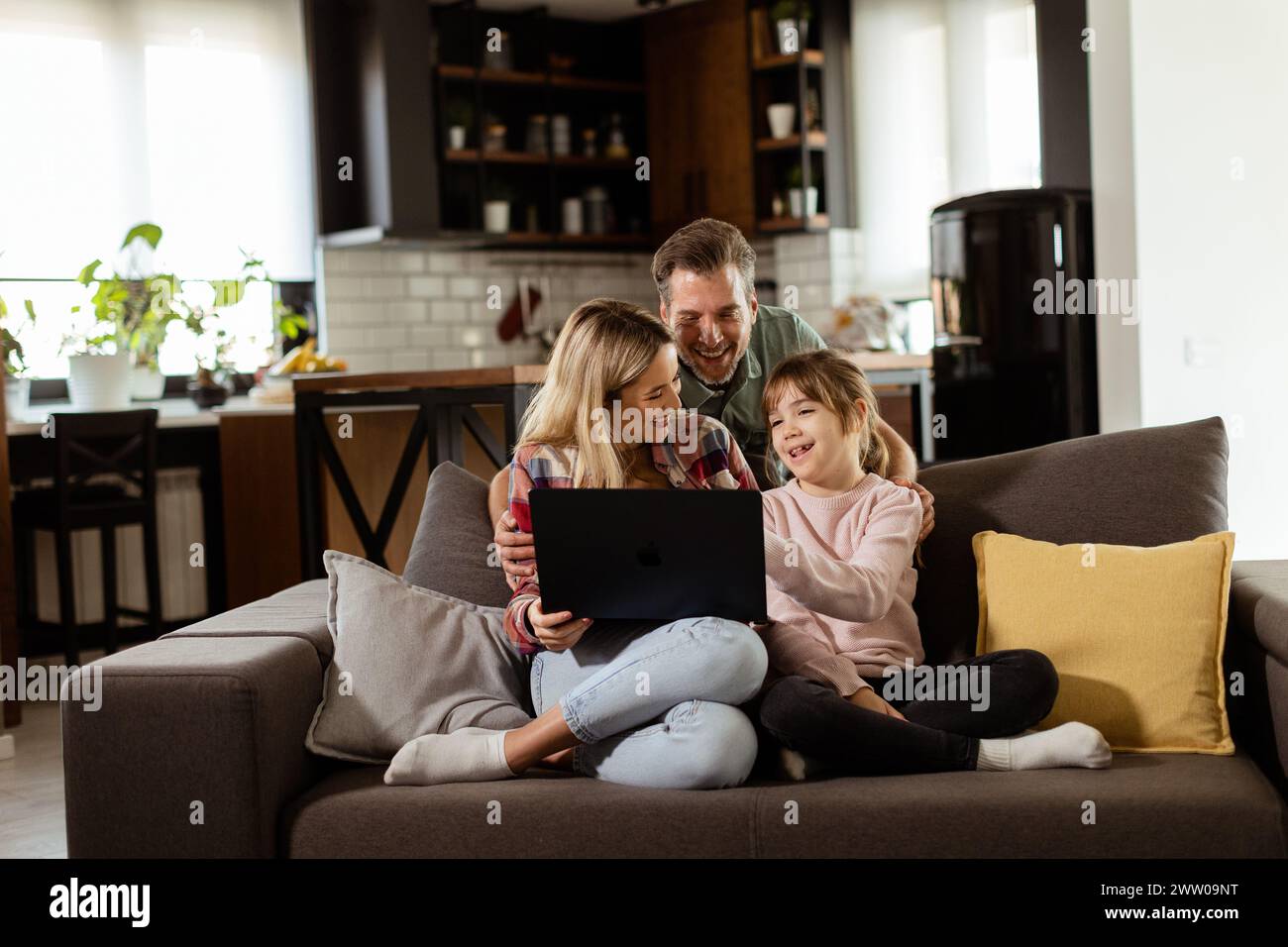Joyful family of three spends quality time together on the living room sofa, sharing a moment around a laptop in their comfortable home Stock Photo