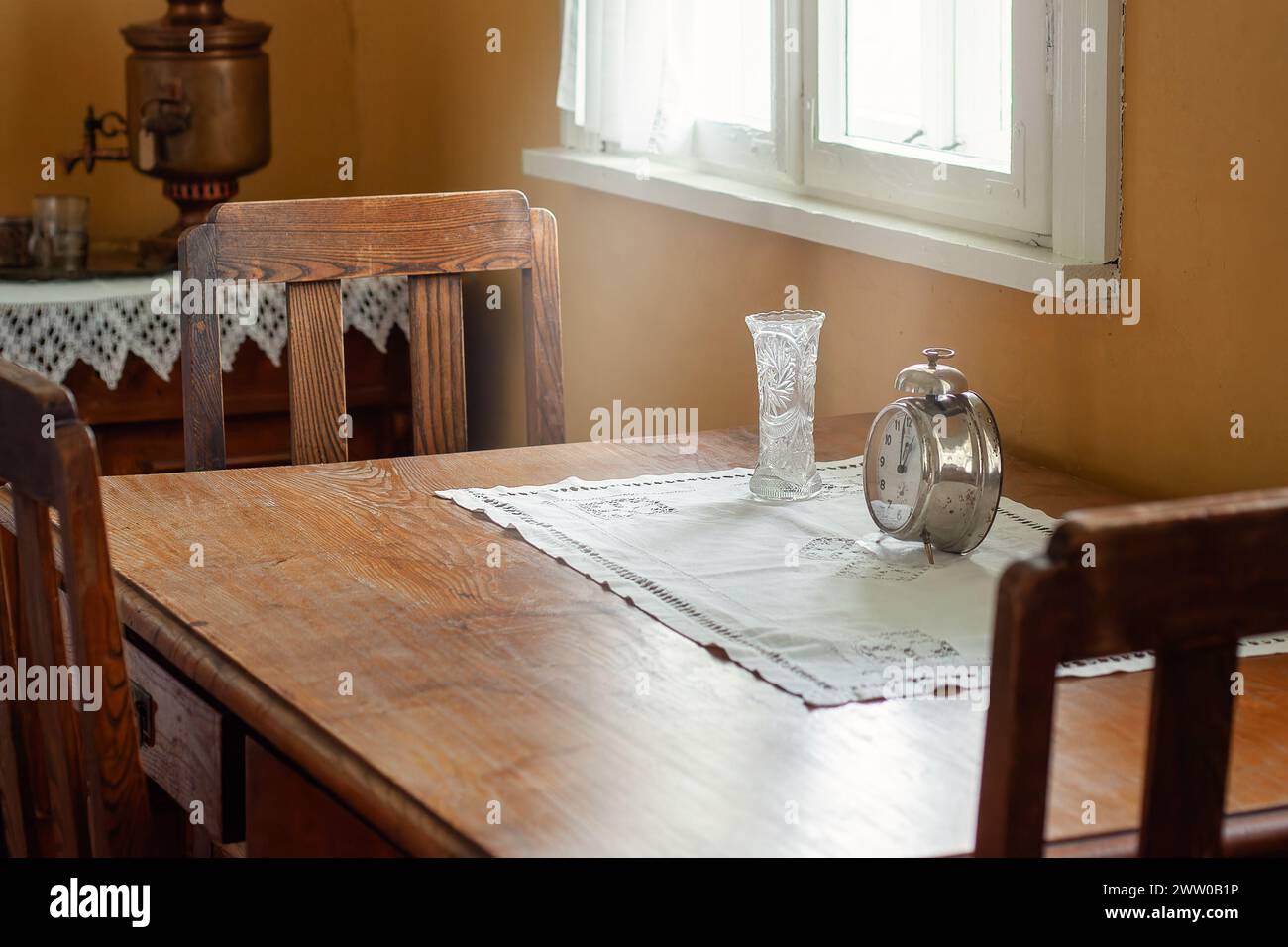 An old oak table and three chairs stand in an antique interior by a window. A white tablecloth, a crystal vase and a large retro alarm clock are place Stock Photo