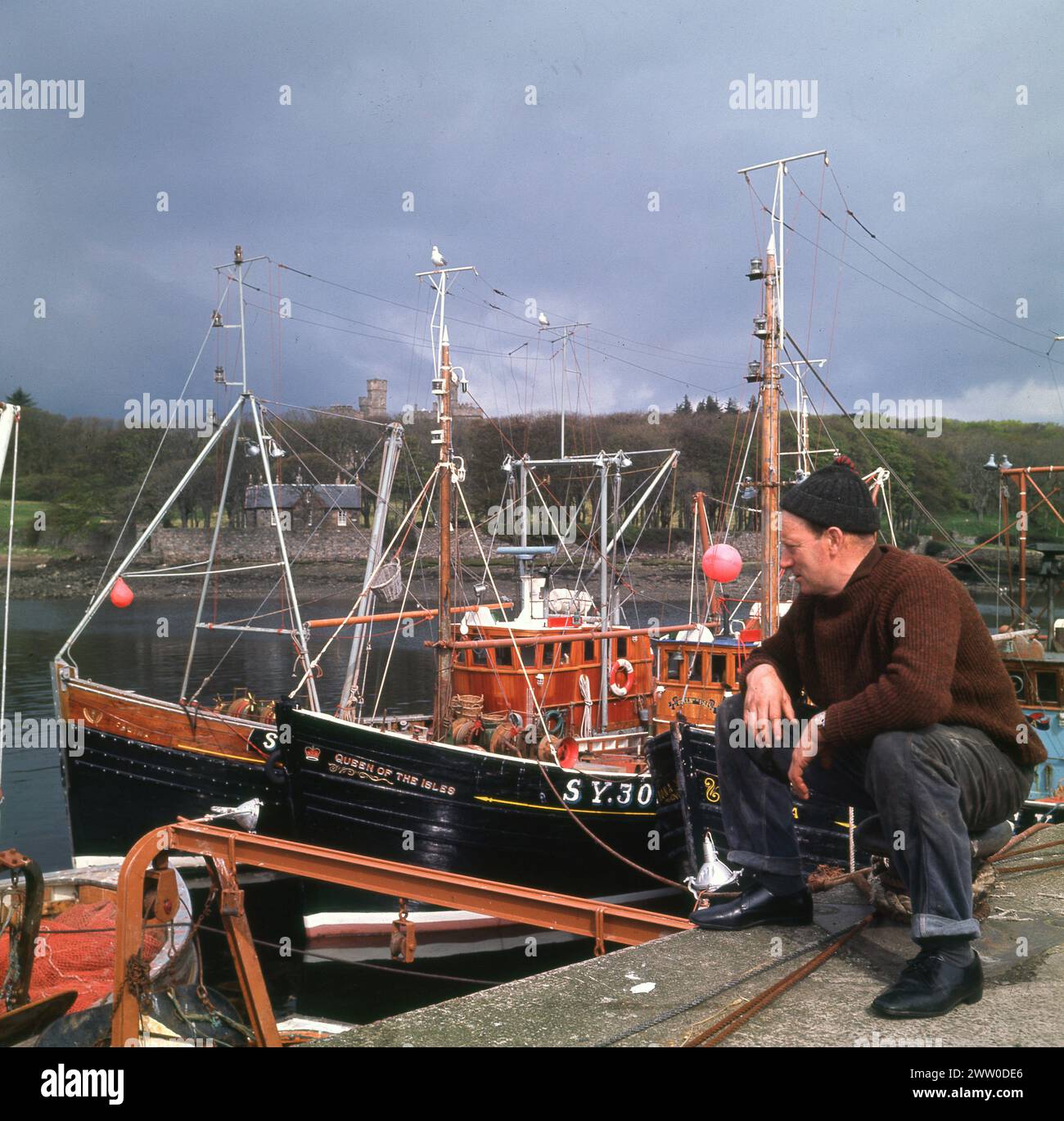 1960s, historical, a fisherman sitting on quay fishing boats, Stornoway, isle of Lewis, Western Isles, Scotland, UK. Stornoway has been the centre of the British herring industry for centuries, with the height of the industry being between 1900 and WW1. While national eating habits saw herring fishing decline, the industry remains economically important for the island. Stock Photo