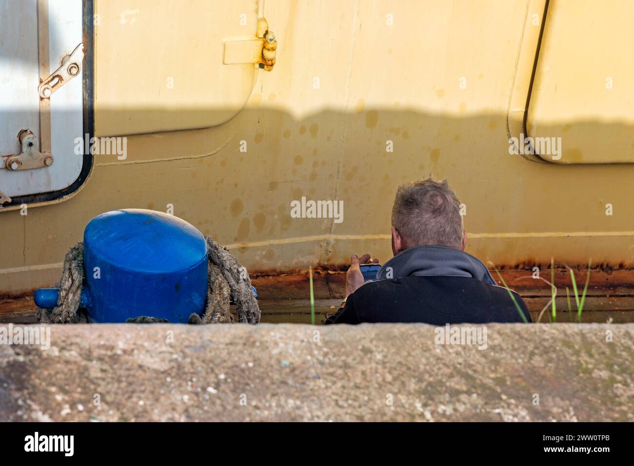 male person photographed from behind is looking at his cell phone with a cigarette in his hand sitting on the deck of a ship Stock Photo