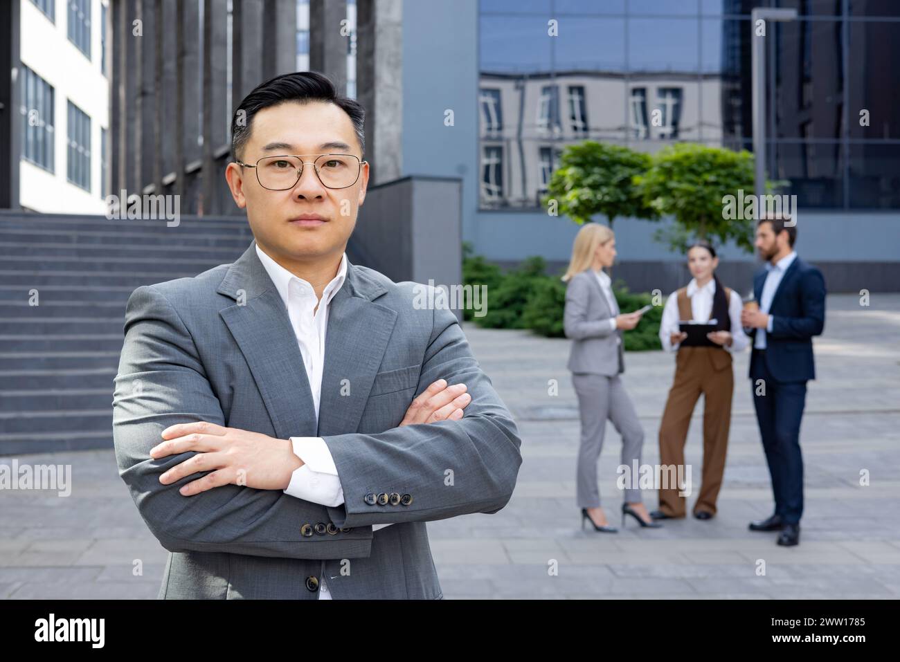 A poised businessman stands with arms crossed as his team discusses in the urban setting, embodying leadership and professionalism. Stock Photo