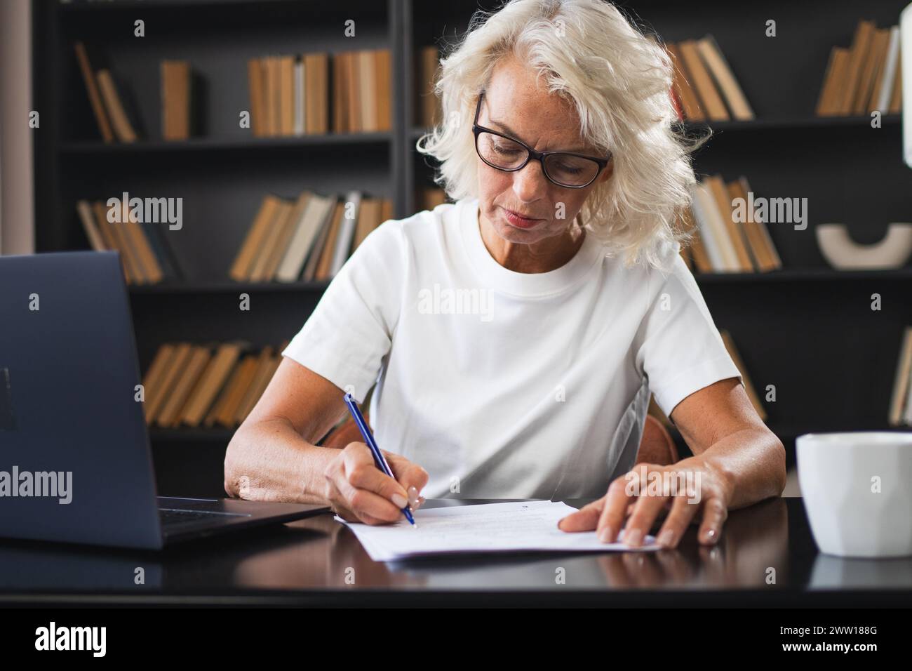 Middle aged senior woman using laptop computer writing notes at workplace. Focused mature old businesswoman work at office making written records doing online work. Mature boss professional worker Stock Photo