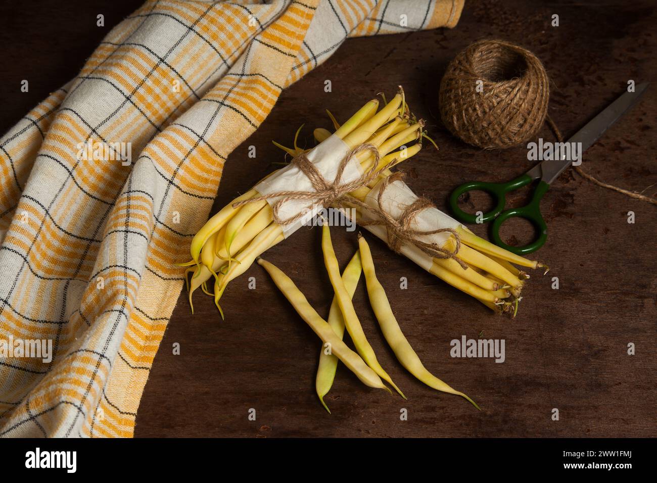 Two bunch and several pods of raw yellow pods of haricot with yellow kitchen towel, scissors and ball of thread on wooden background. French beans or Stock Photo