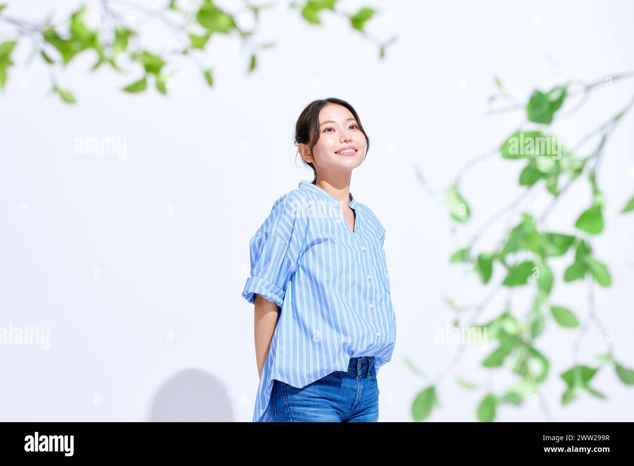 Asian woman in blue shirt and jeans standing in front of green tree Stock Photo