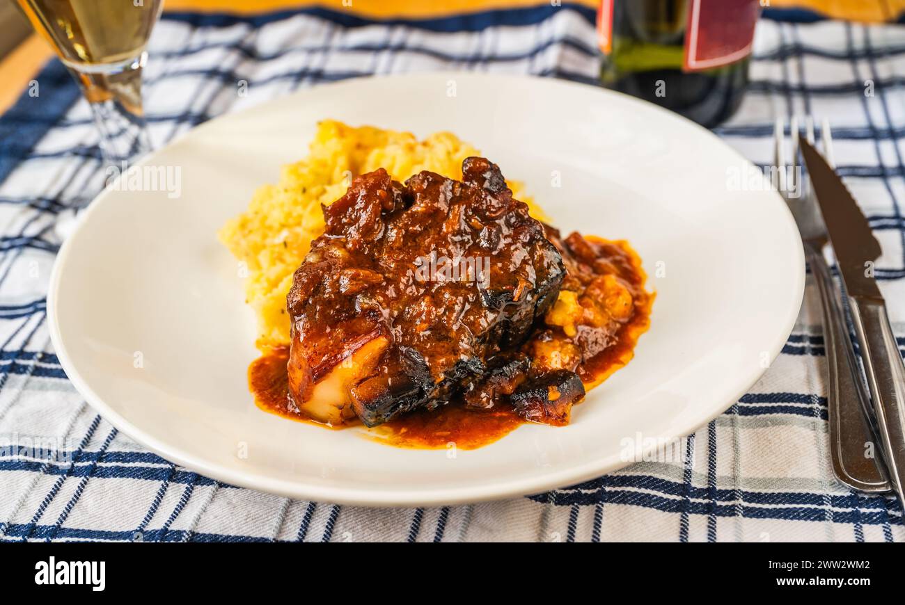 Baked juicy pork rib piece in spicy red sauce with mashed potato on white plate, cutlery and wine on table, closeup. Stock Photo