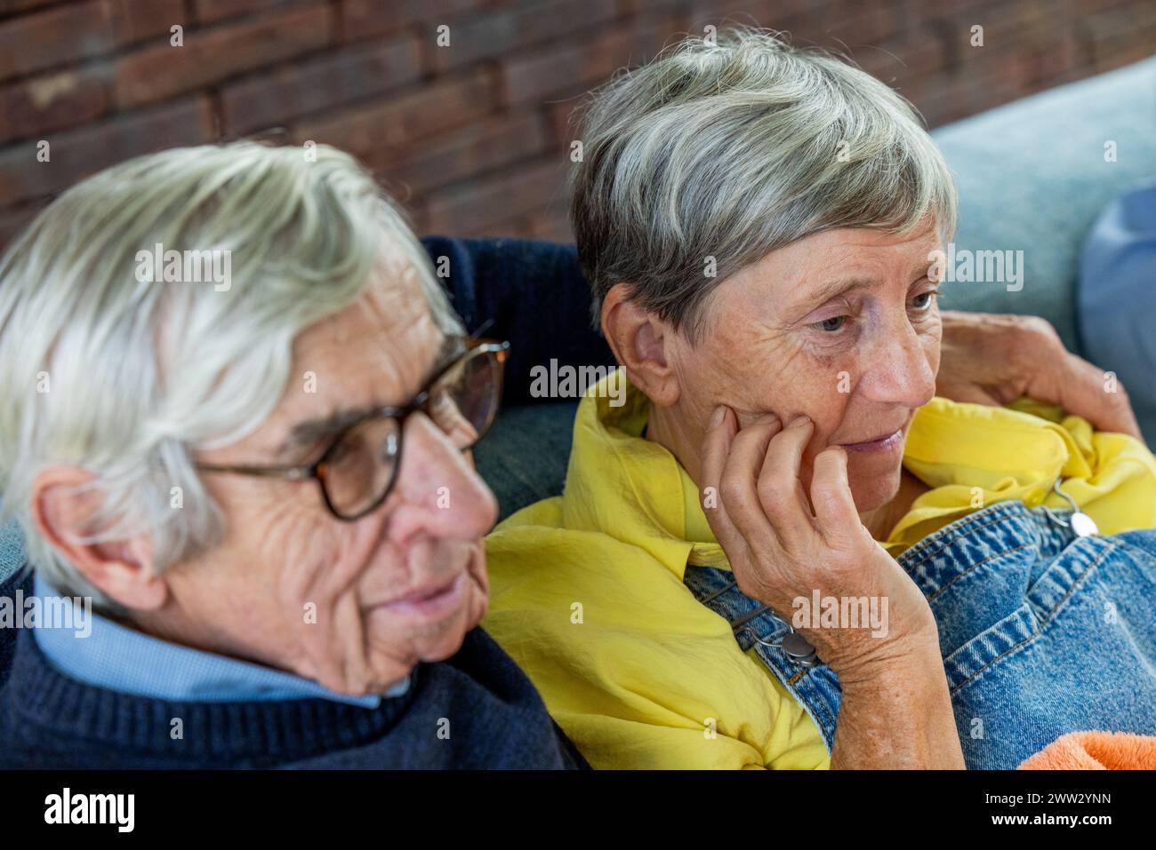 Close-up of a contemplative senior woman with her hand on her cheek, sitting closely next to her male companion. They are both in a thoughtful state, suggesting a moment of reflection or concern, with a cozy indoor backdrop accentuated by a brick wall. Pensive Senior Woman with Companion in Thoughtful Moment. High quality photo Stock Photo