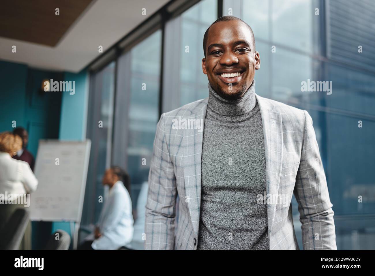 Young African male entrepreneur stands confidently in a modern office boardroom, surrounded by a diverse and professional team. Smiling at the camera, Stock Photo