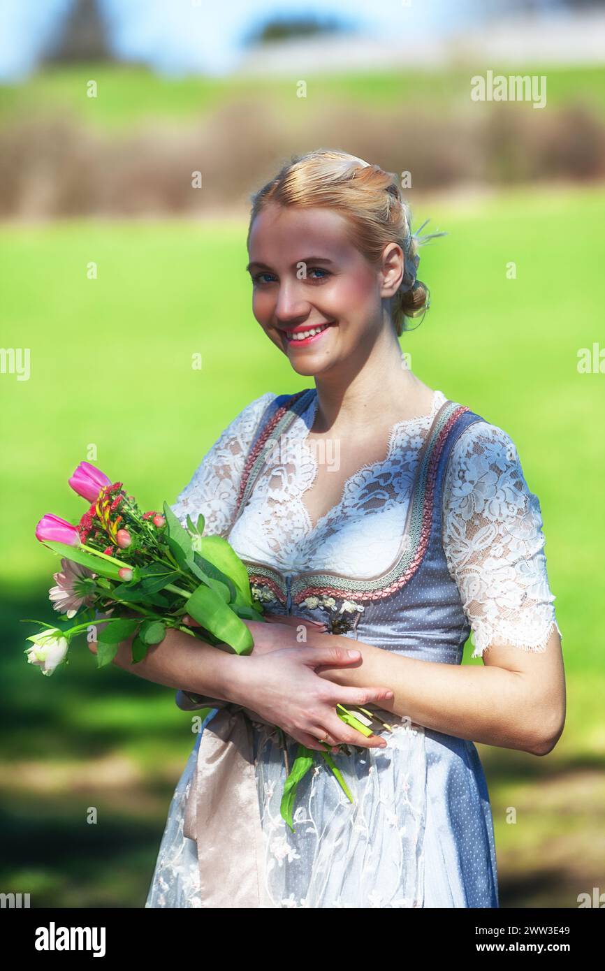 A Smiling Woman With Blonde Hair And A Dirndl Stands In A Meadow, Holding Tulips And Looking Into The Camera. 3/4 Shot. Stock Photo