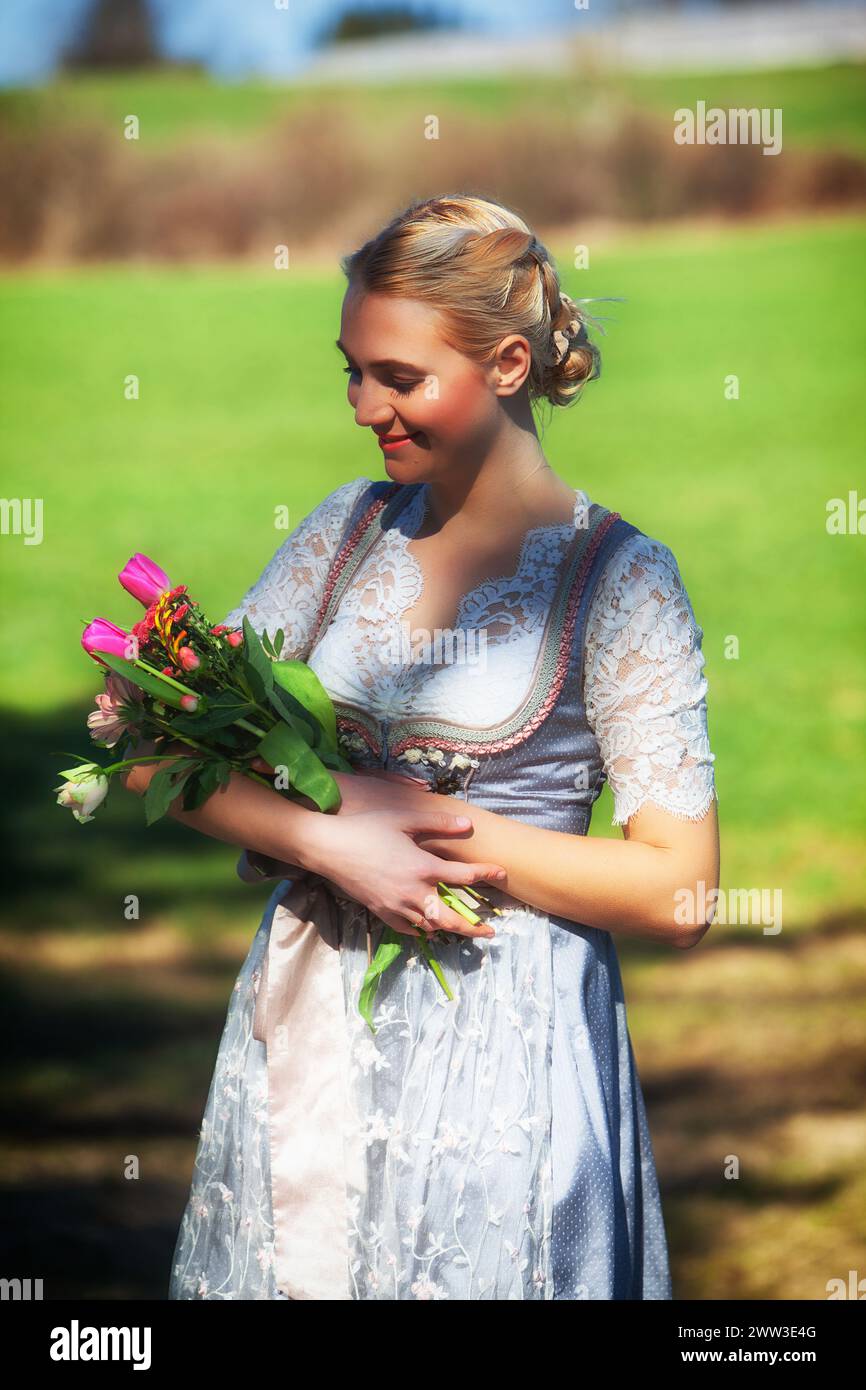 A Young Woman With Blonde Hair And A Dirndl Stands Smiling In A Flowering Meadow, Holding Tulips In Her Arms. Stock Photo