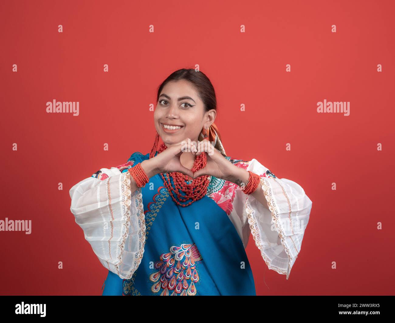 latina girl in ecuadorian indian costume happy making a heart sign with her hands on a red background Stock Photo