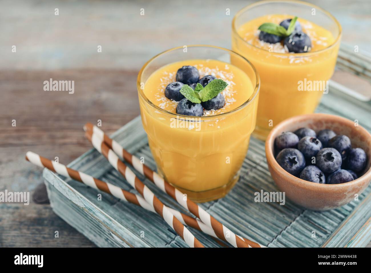 Refreshing and healthy mango smoothie in glasses with coconut flakes and fresh blueberries on wooden background Stock Photo