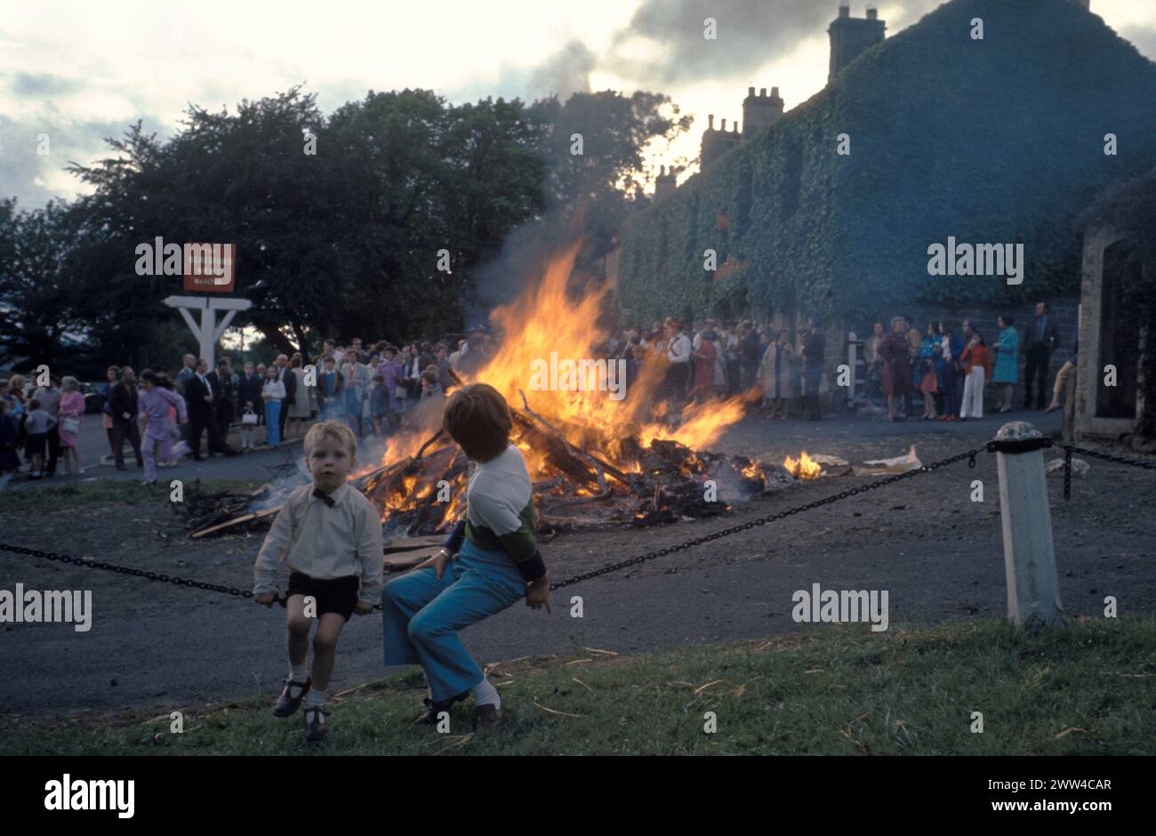 Baal Fire Whalton Northumberland UK. St Johns Eve 23 June annually, outside Beresford Arms in centre of village. 1971 or 1972 The tradition was to take a lighted ember home ensure good luck the following year. Crowd of people and kids in the centre of village watch the fire die down. 1970 UK HOMER SYKES Stock Photo