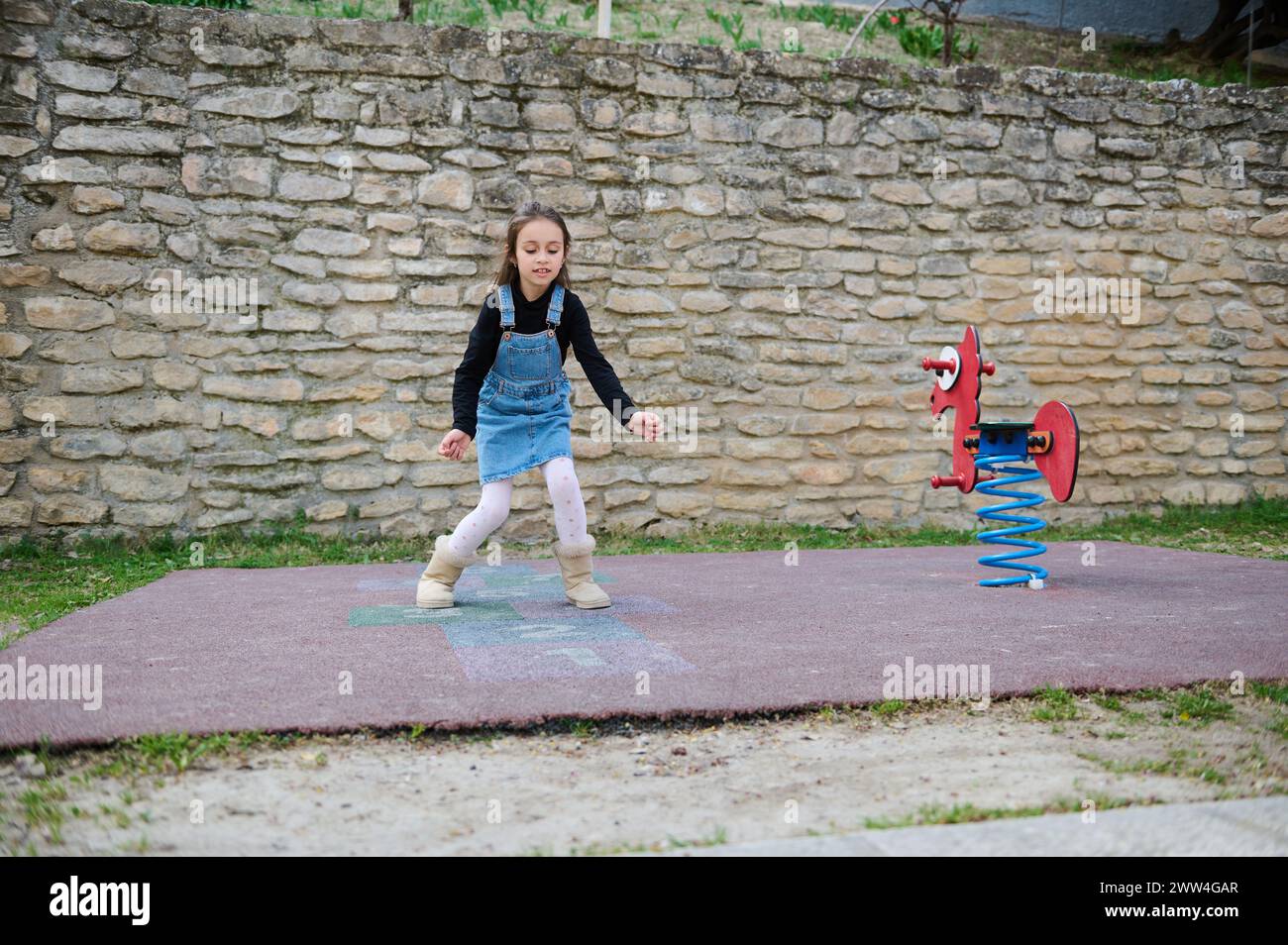 Active little child girl, elementary age schoolgirl plays hopscotch, takes turns jumping over squares marked on the ground. Street children's games in Stock Photo