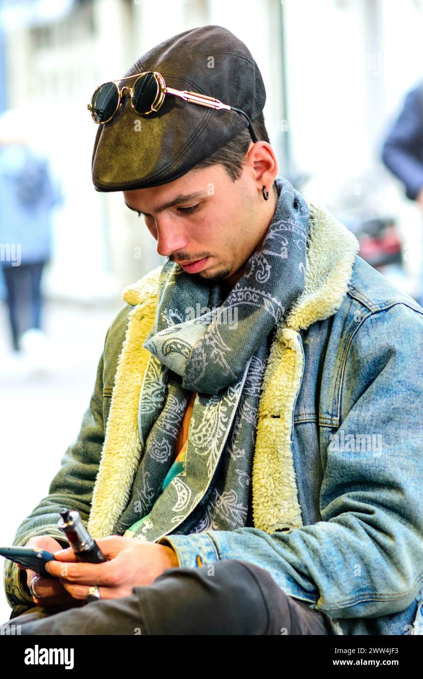 Young man in denim jacket sitting at outdoor table studying smartphone - Tours, Indre-et-Loire (37), France. Stock Photo