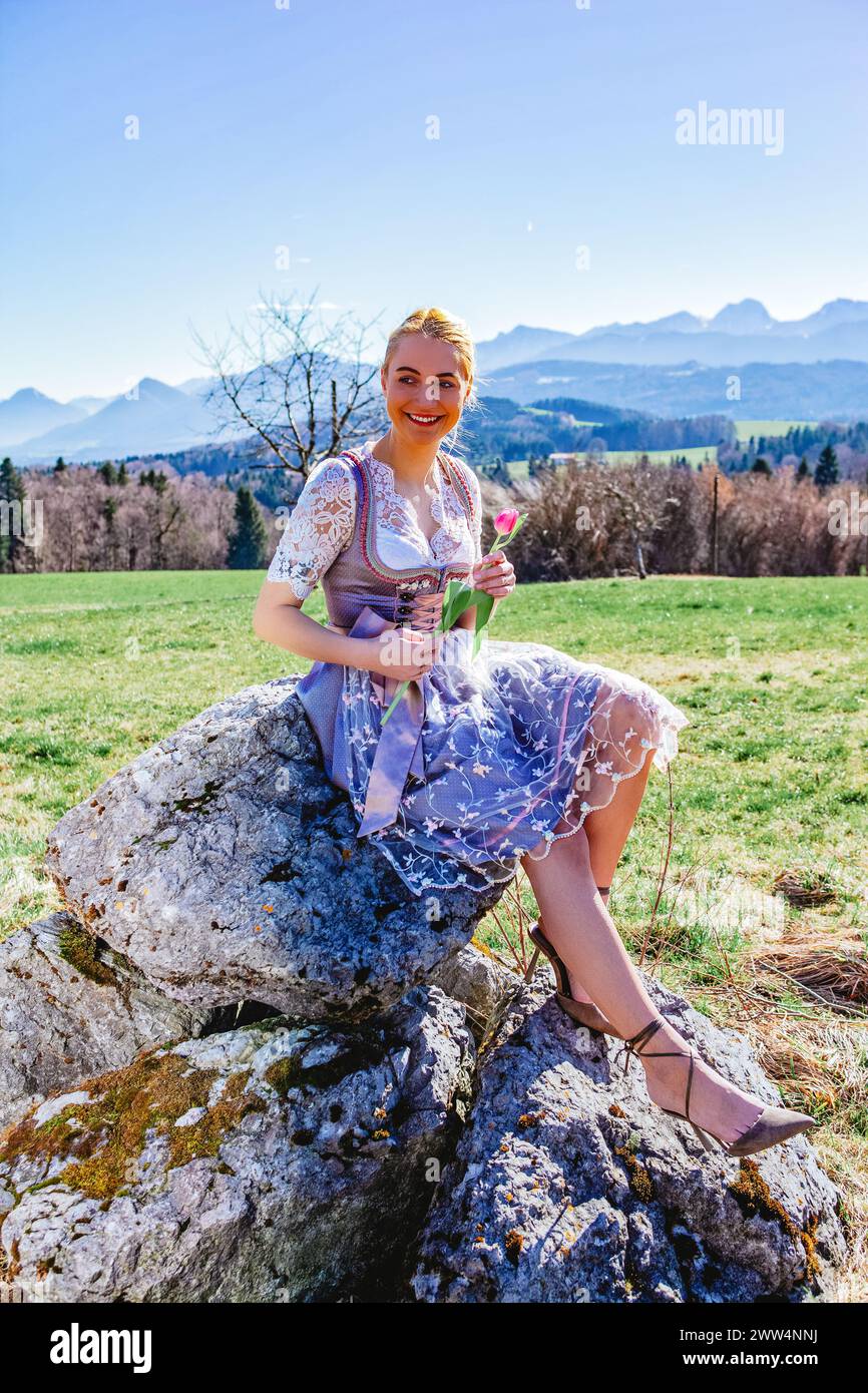 A Woman With Blonde Hair And A Dirndl Sits Smiling On A Stone In Front Of Snow-covered Mountains, Holding A Tulip And Beaming At The Camera. Stock Photo