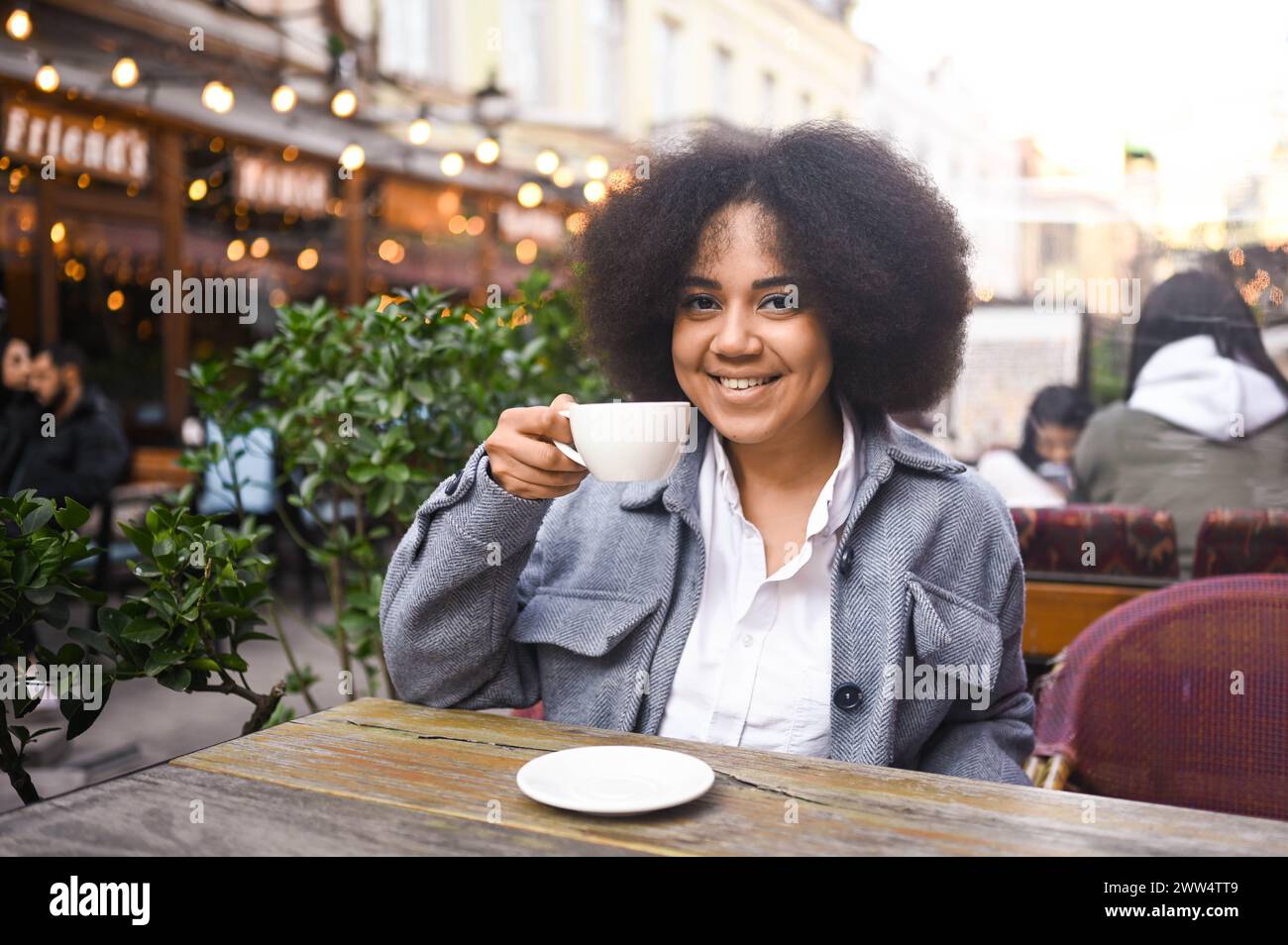 Fashion street style portrait of attractive young natural beauty African American woman with afro hair in tweed jacket posing outdoors in sidewalk cafe. Happy lady with coffee cup in big city Stock Photo